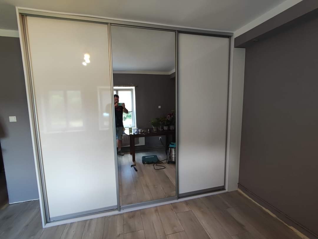 Biała szafa wnękowa z przesuwnymi drzwiami, z lustrzanym środkowym panelem, odbijającym fragment pokoju z drewnianym stołem i osobą robiącą zdjęcie.