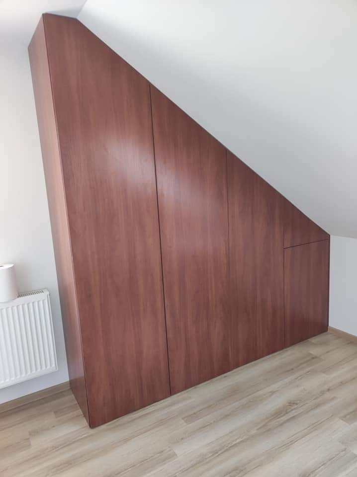 Brązowa szafa z drewnianymi frontami, dopasowana do skosu poddasza, z widocznym grzejnikiem i fragmentem podłogi imitującej drewno.