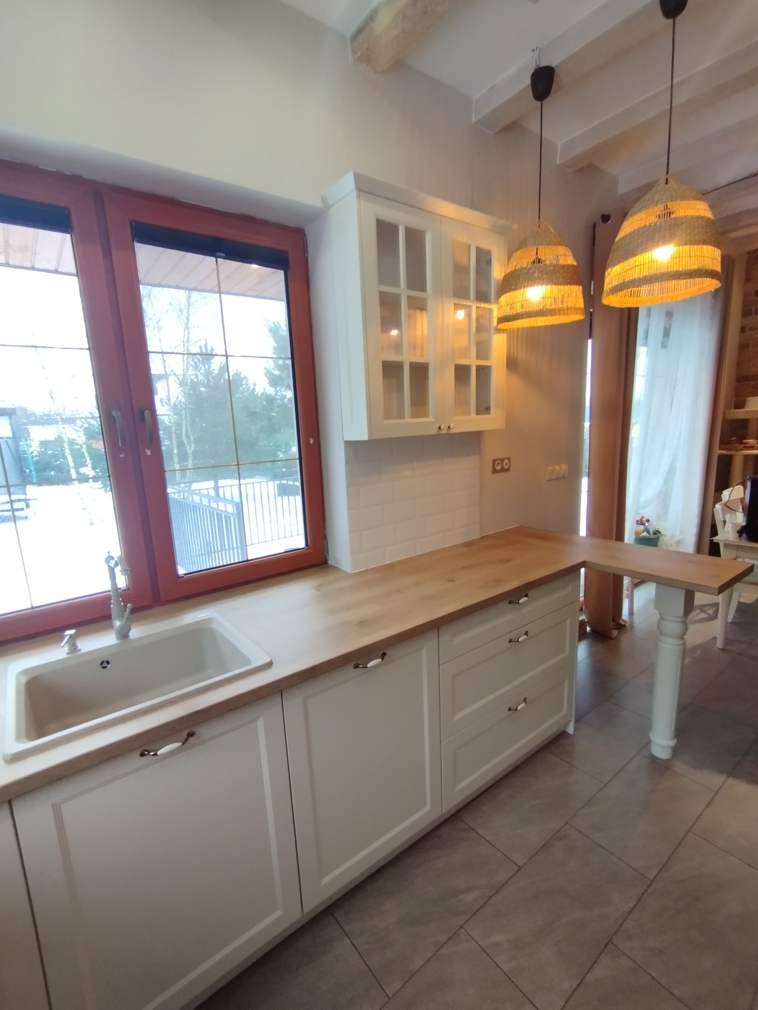 Jasna kuchnia z drewnianym blatem, białymi szafkami z frezowanymi frontami i złotymi uchwytami, widok na okno z widokiem na zimowy krajobraz, oświetlona trzema plecionymi lampami.