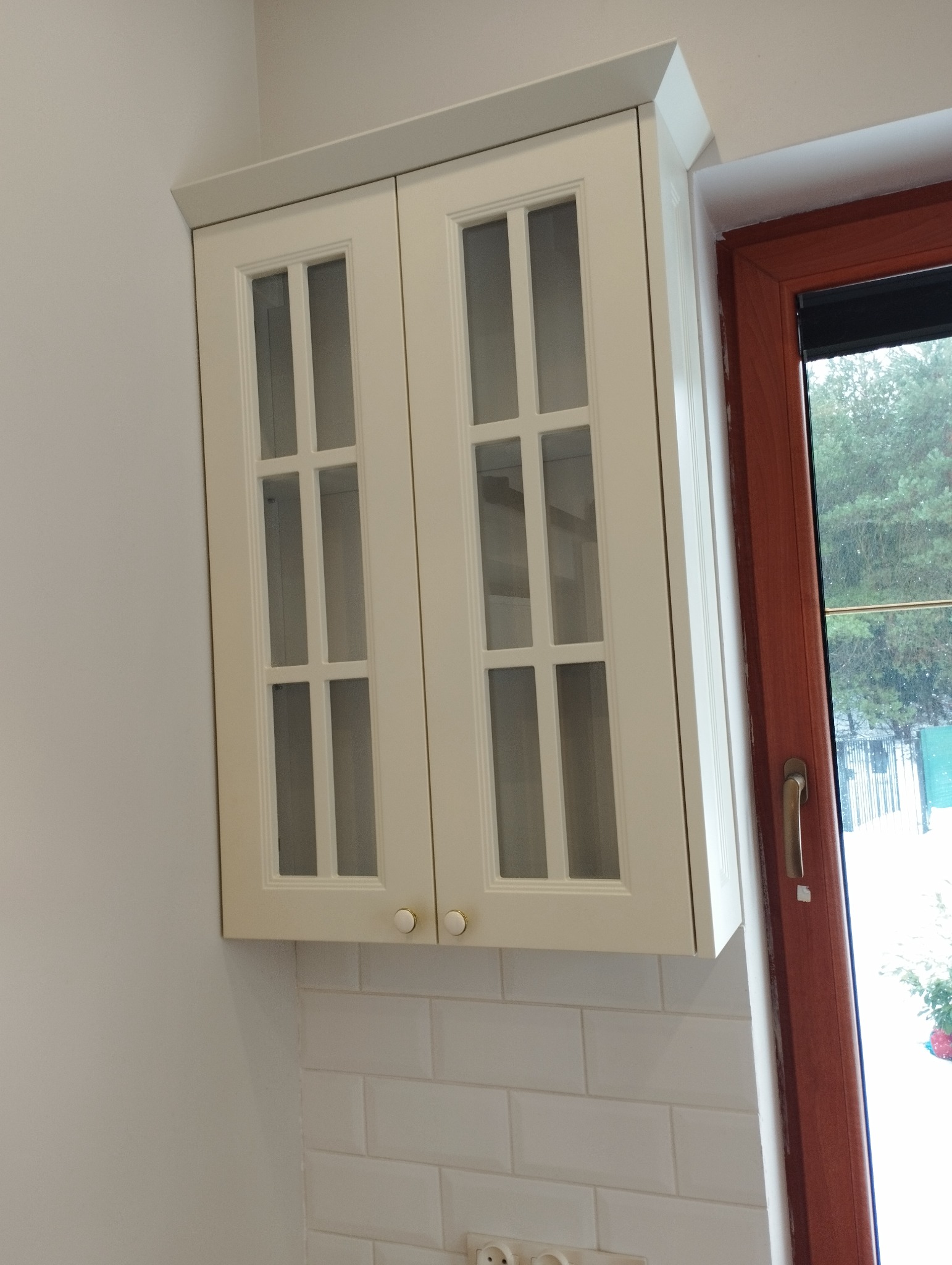 Kremowa szafka kuchenna z przeszklonymi drzwiczkami, zamontowana nad białą ścianą z płytek typu metro, obok okna z brązową ramą.