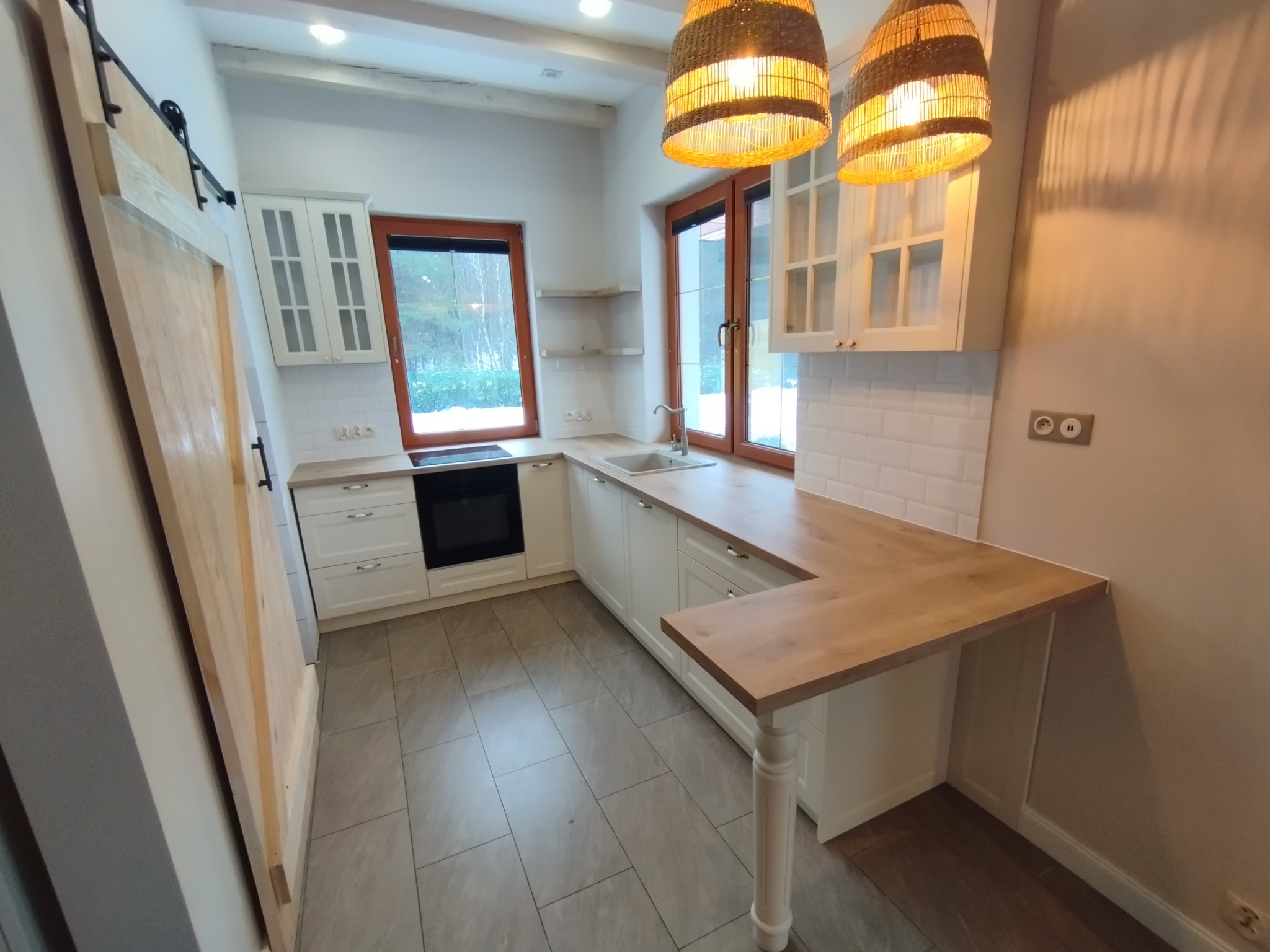 Jasna kuchnia z białymi szafkami i drewnianym blatem, oświetlona dwoma wiklinowymi lampami, z przesuwnymi drzwiami do spiżarni po lewej stronie i widokiem za oknem na zewnątrz.