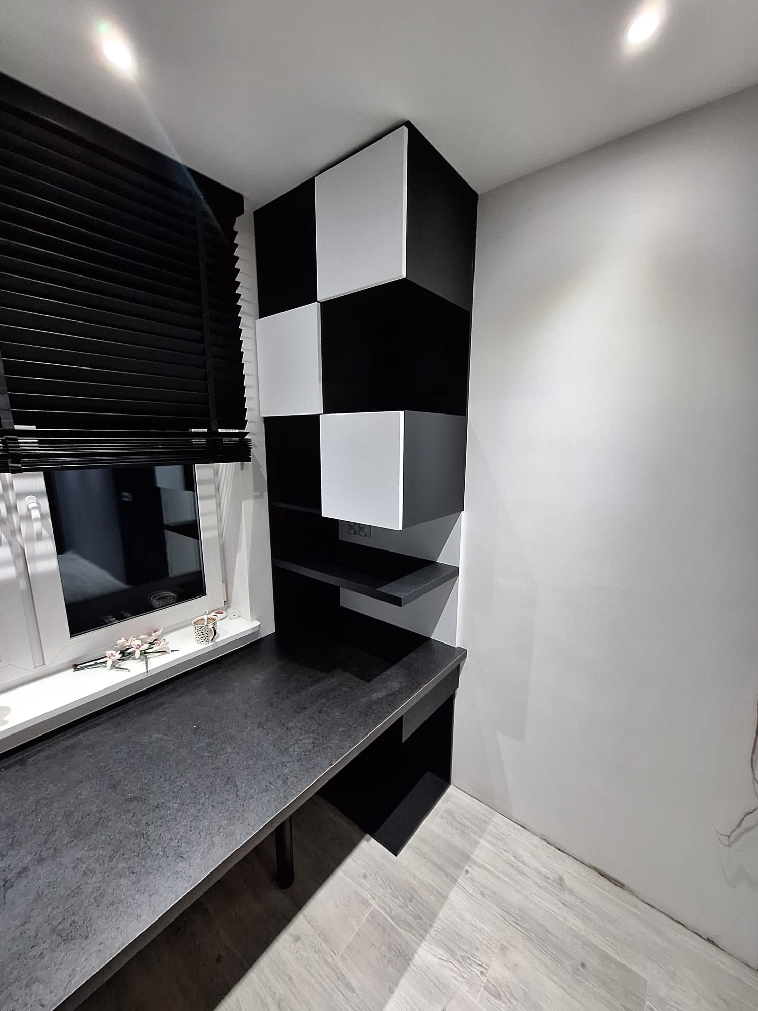 Czarno-biała zabudowa biurka z półkami w szachownicę, zintegrowana z blatem roboczym przy oknie z żaluzjami.