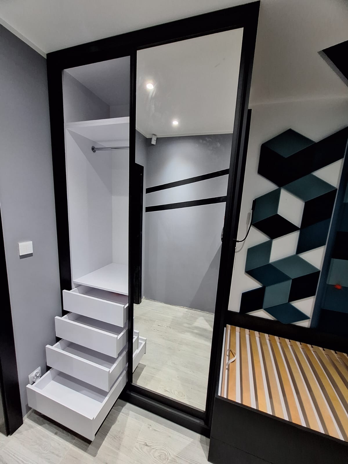 Nowoczesna szafa wnękowa z lustrzanymi drzwiami przesuwnymi, widoczne białe szuflady i półka, czarna rama, na ścianie dekoracja z geometrycznych brył.