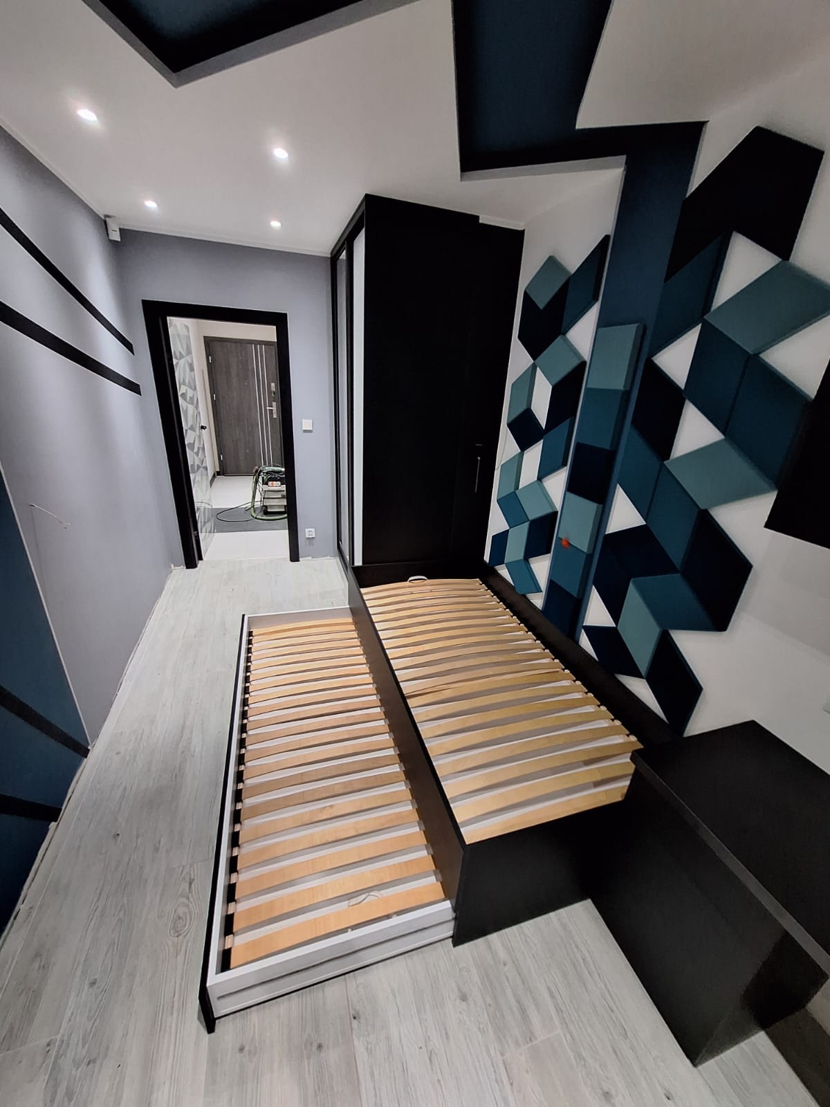 Wysuwane łóżko z drewnianym stelażem w nowoczesnej sypialni z geometryczną dekoracją ścienną w odcieniach błękitu i czerni, widok na korytarz.