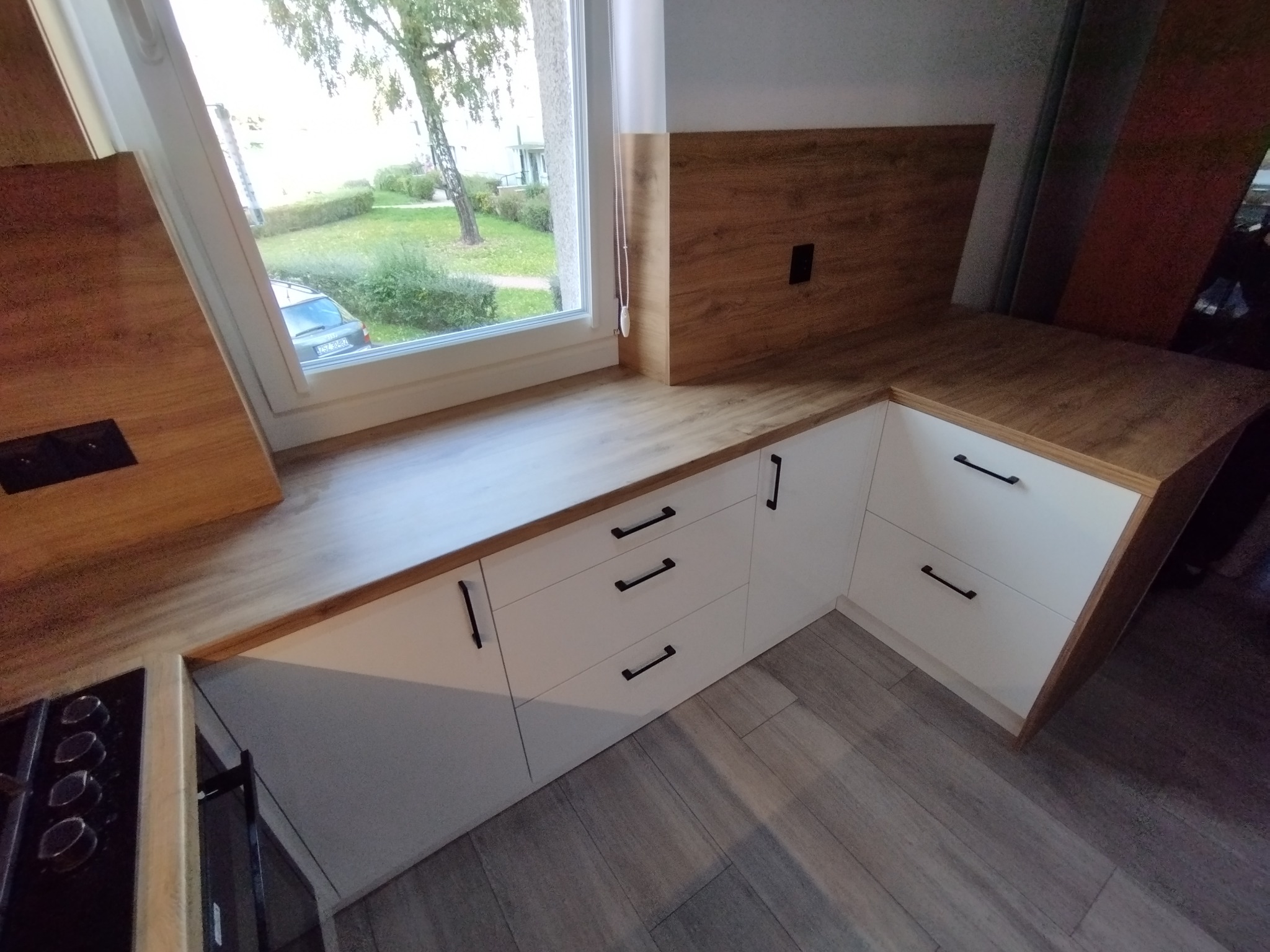 Fragment białej zabudowy kuchennej z drewnianym blatem i czarnymi uchwytami, widok z góry na szuflady i blat pod oknem z widokiem na zieleń.