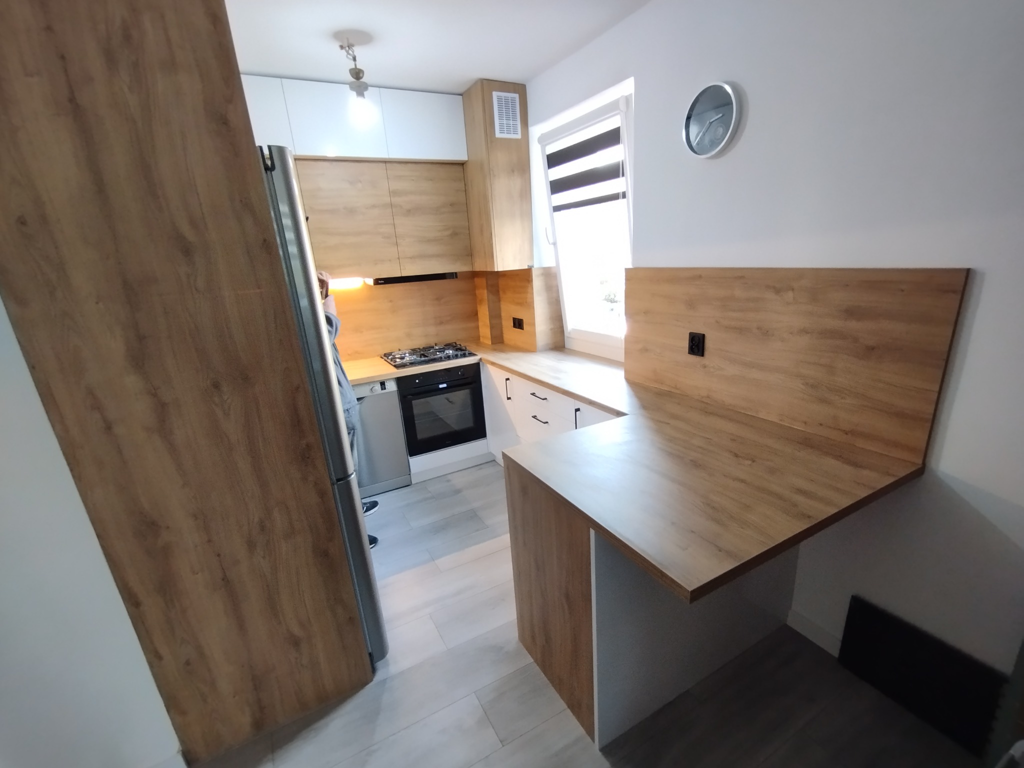 Nowoczesna kuchnia z drewnianymi frontami i blatem, widok z boku, z elementami białymi, srebrny zegar na ścianie, wąskie okno z roletami.