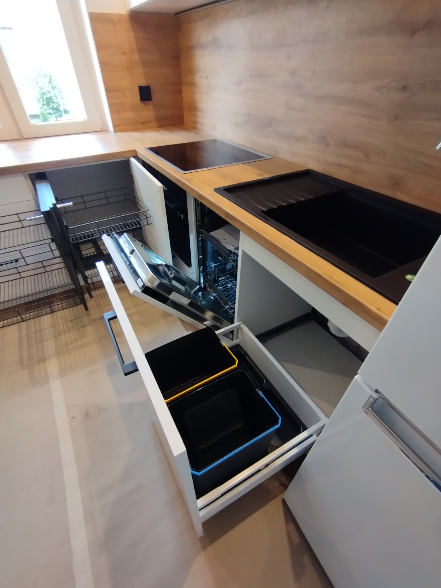 Nowoczesna kuchnia z drewnianym blatem, czarnym zlewem i otwartymi szufladami: kosz na śmieci z niebieską obwódką i wysuwany kosz cargo.