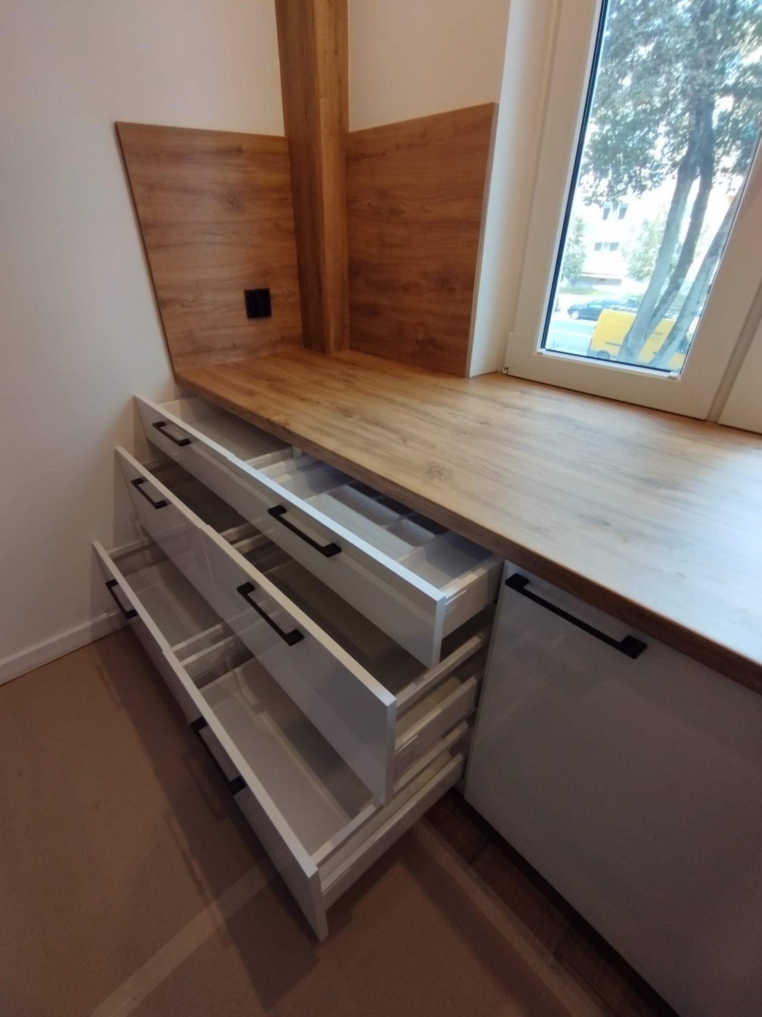 Białe szuflady z czarnymi uchwytami, częściowo wysunięte, zamontowane pod drewnianym blatem przy oknie z widokiem na drzewa i ulicę.