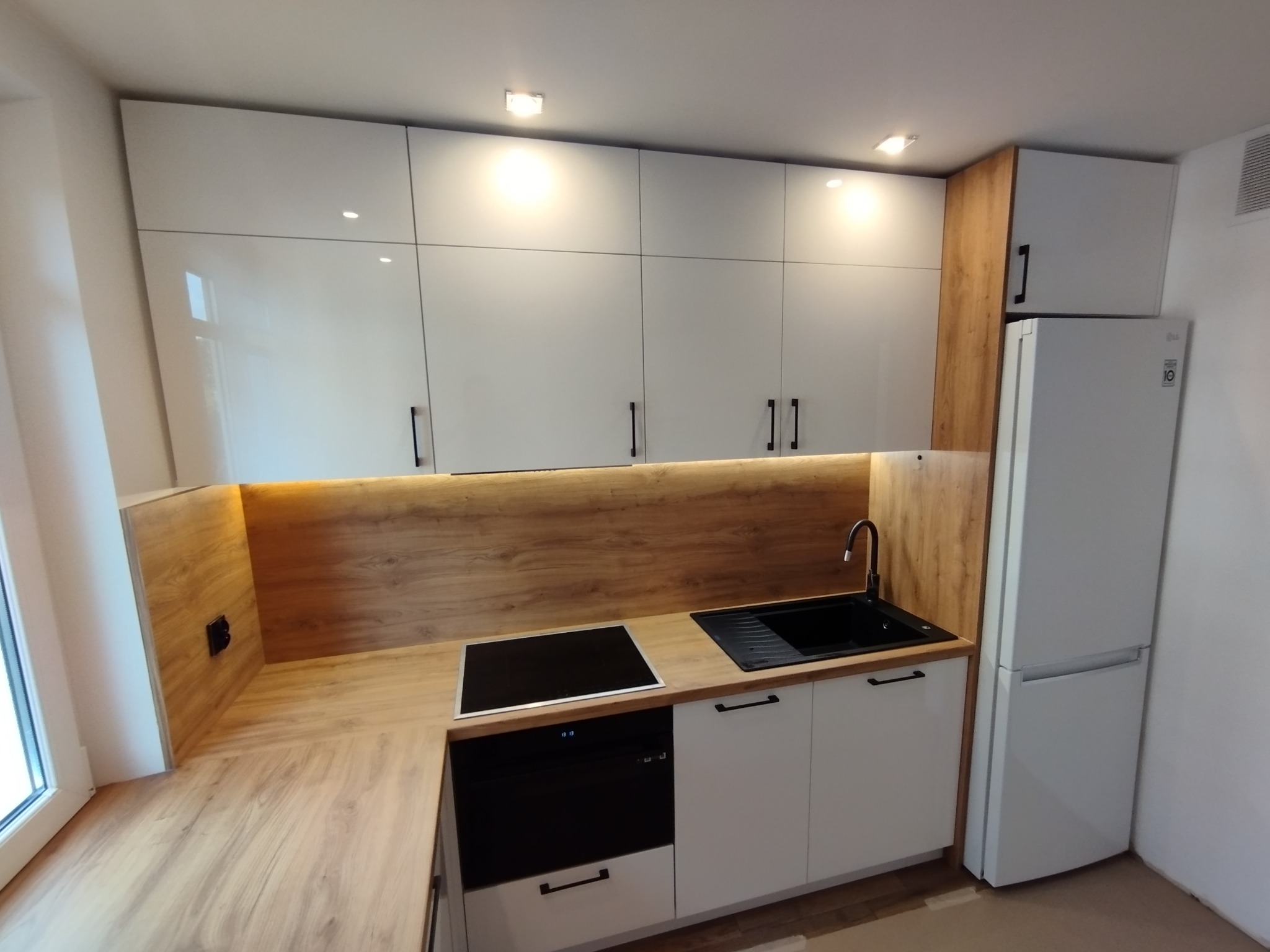 Nowoczesna kuchnia z białymi szafkami, drewnianym blatem i czarnymi akcentami, w tym zlewozmywakiem i płytą grzewczą, oświetlona ciepłym światłem LED pod szafkami wiszącymi.