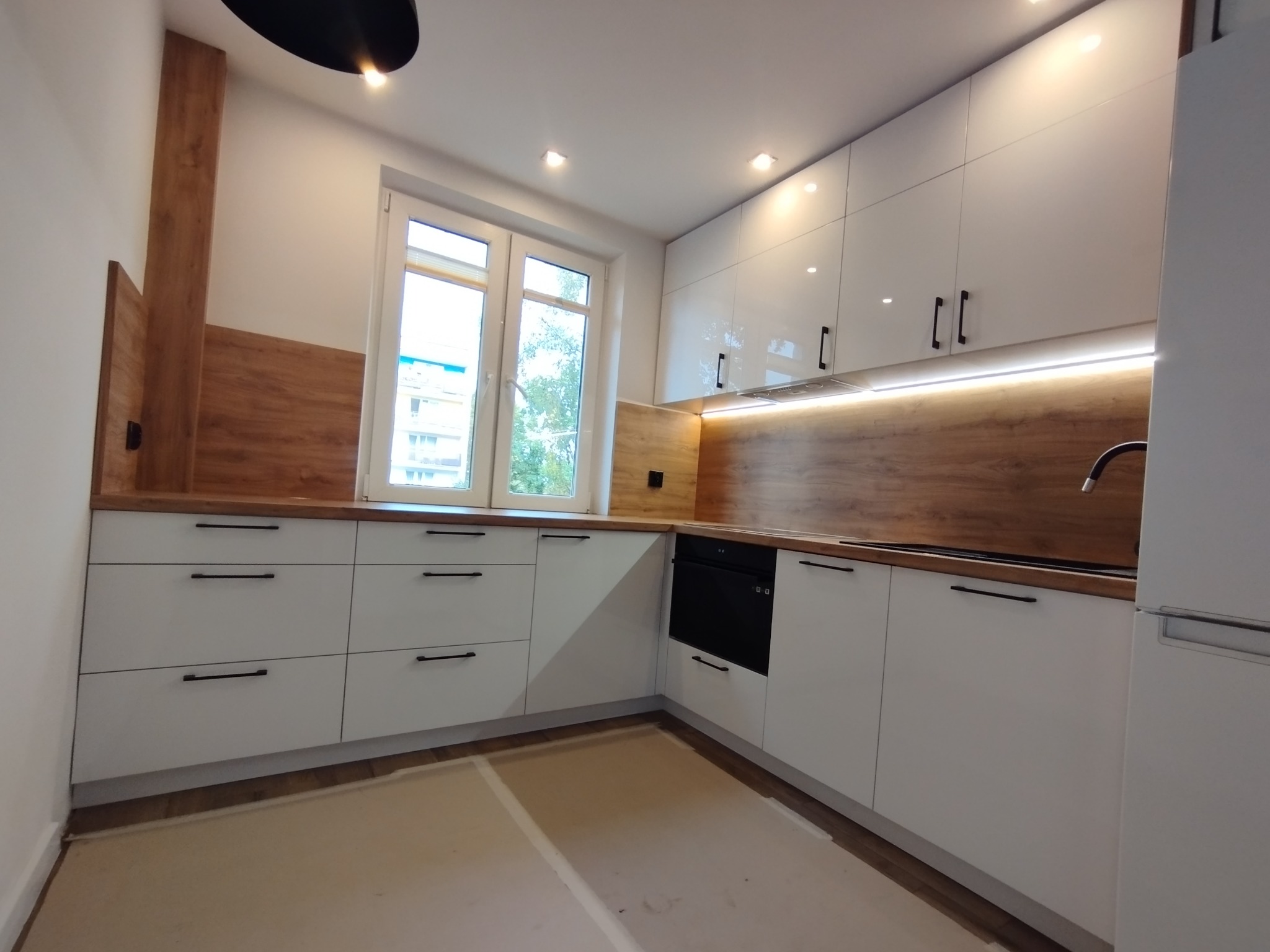 Nowoczesna, jasna kuchnia z białymi, lakierowanymi frontami szafek i drewnianym blatem, oświetlona listwą LED pod szafkami wiszącymi. Widoczne okno z widokiem na budynek mieszkalny.