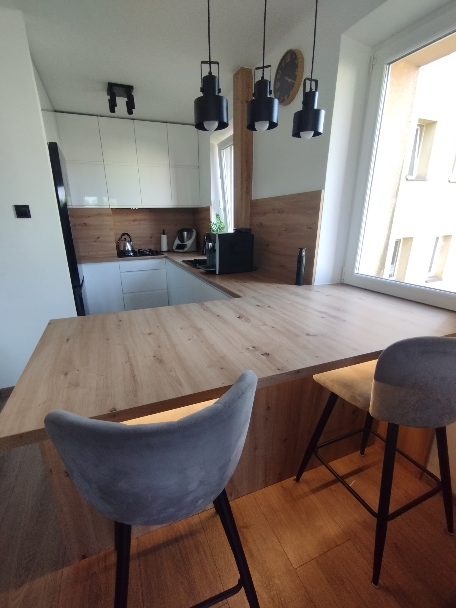 Nowoczesna kuchnia z białymi szafkami, drewnianym blatem i wyspą z dwoma szarymi hokerami, oświetlona trzema czarnymi lampami wiszącymi.