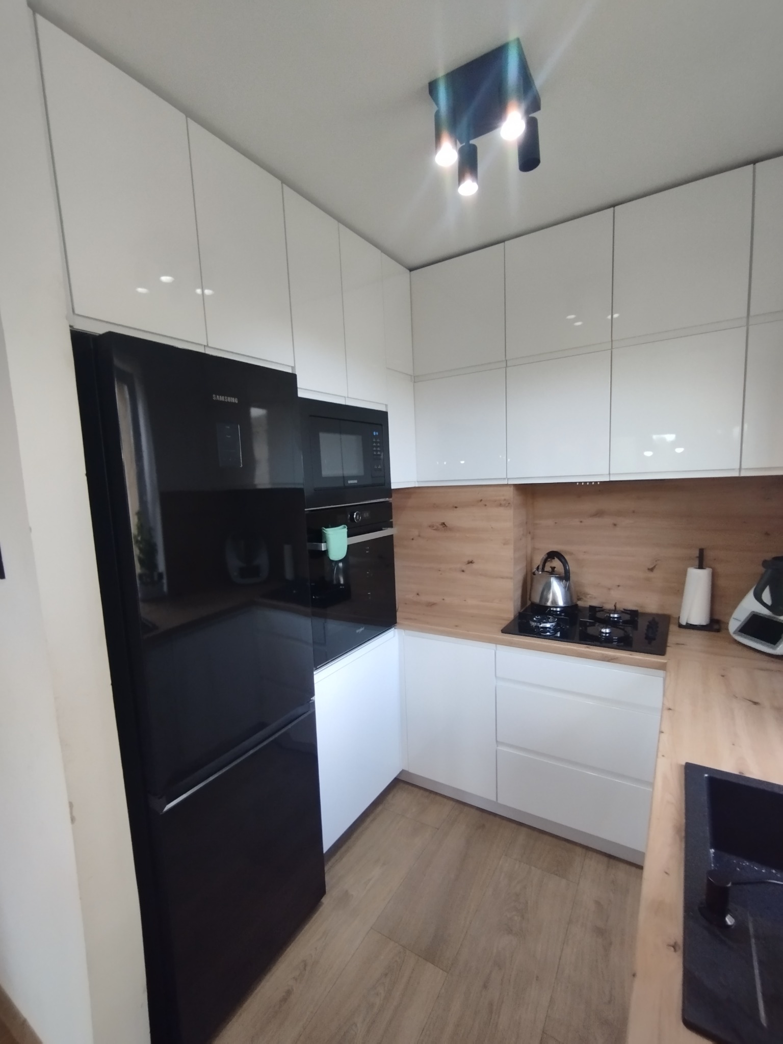 Nowoczesna, jasna kuchnia w Jeleninie z białymi, błyszczącymi szafkami, drewnianym blatem i czarnym sprzętem AGD, w tym lodówką Samsung i płytą grzewczą.