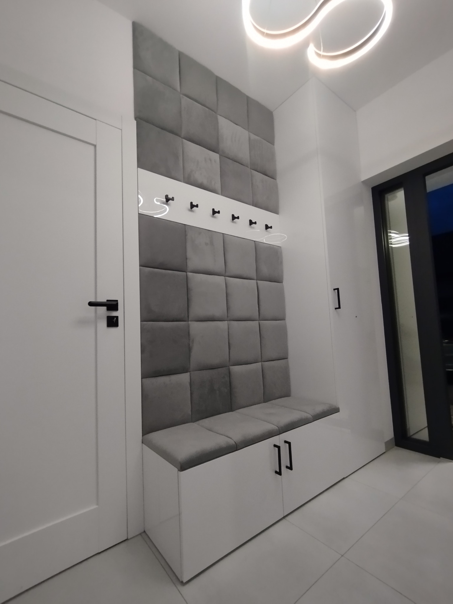 Nowoczesna zabudowa przedpokoju z siedziskiem i panelem tapicerowanym w odcieniach szarości, uzupełniona o wieszaki i białe fronty szafek, widoczna obok białych drzwi i minimalistycznego...