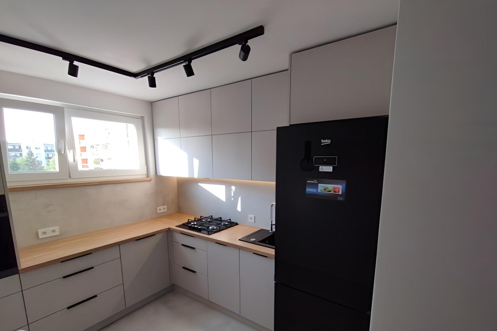Jasnoszara zabudowa kuchenna z drewnianym blatem, czarną lodówką i oświetleniem szynowym punktowym