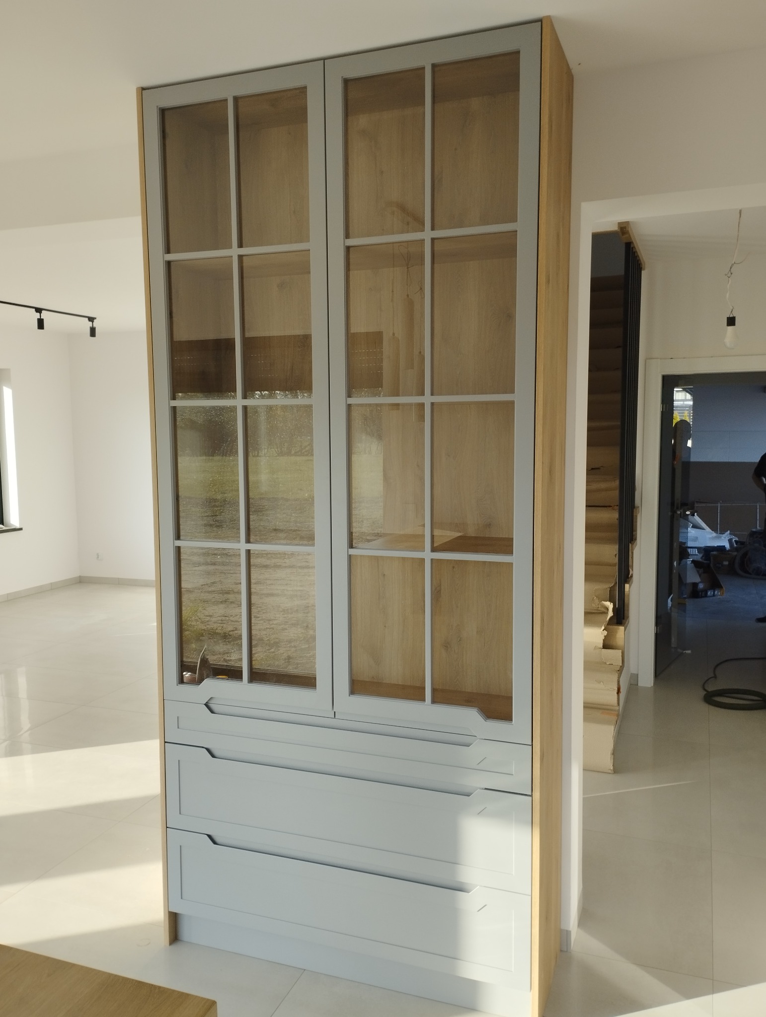 Narożna szafa z przeszklonymi drzwiczkami w szarych ramach i trzema szufladami w jasnym odcieniu szarości, z widoczną strukturą drewna na bokach, umieszczona w nowoczesnym wnętrzu z białymi...