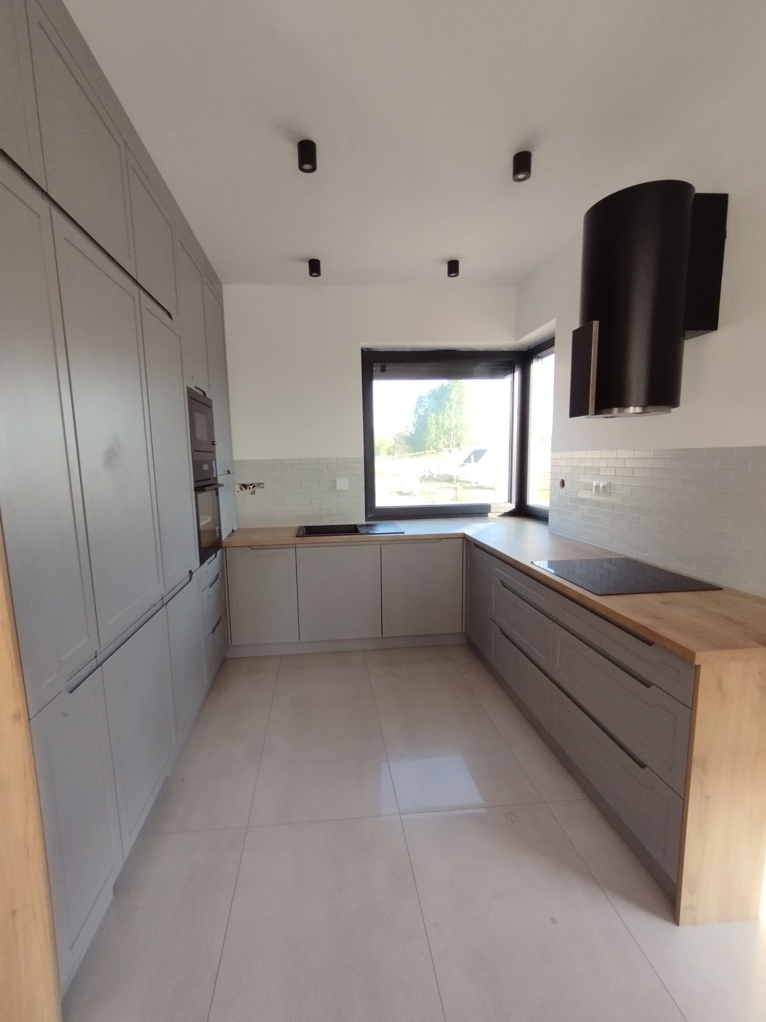 Jasna kuchnia w szarościach z drewnianymi blatami, minimalistyczna zabudowa z piekarnikiem w słupku, czarny okap i punktowe oświetlenie sufitowe, widok na zewnątrz przez duże okno.