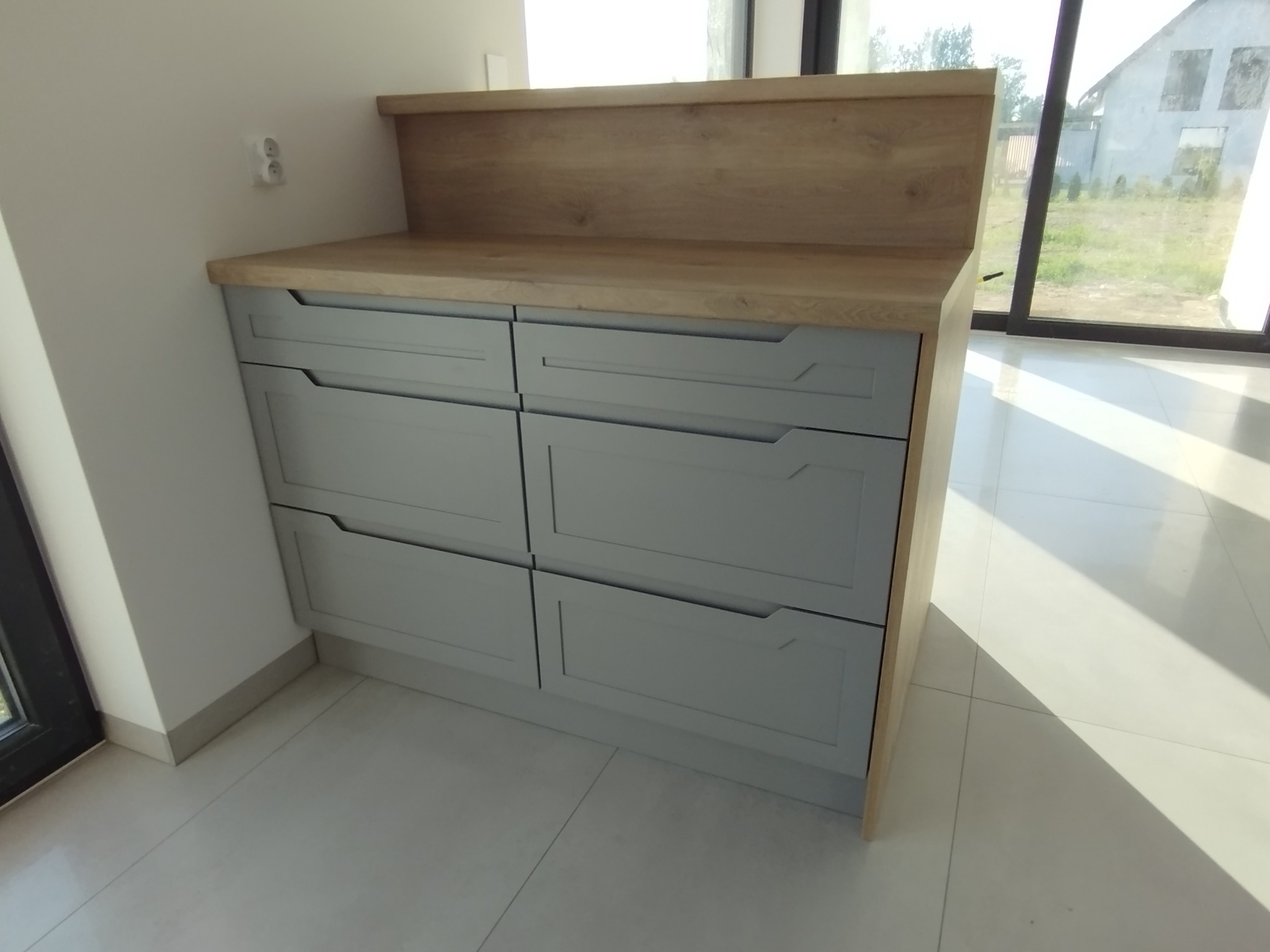 Szara komoda z drewnianym blatem i nadstawką, nowoczesny design, minimalistyczne uchwyty, jasne płytki podłogowe, duże okno w tle.