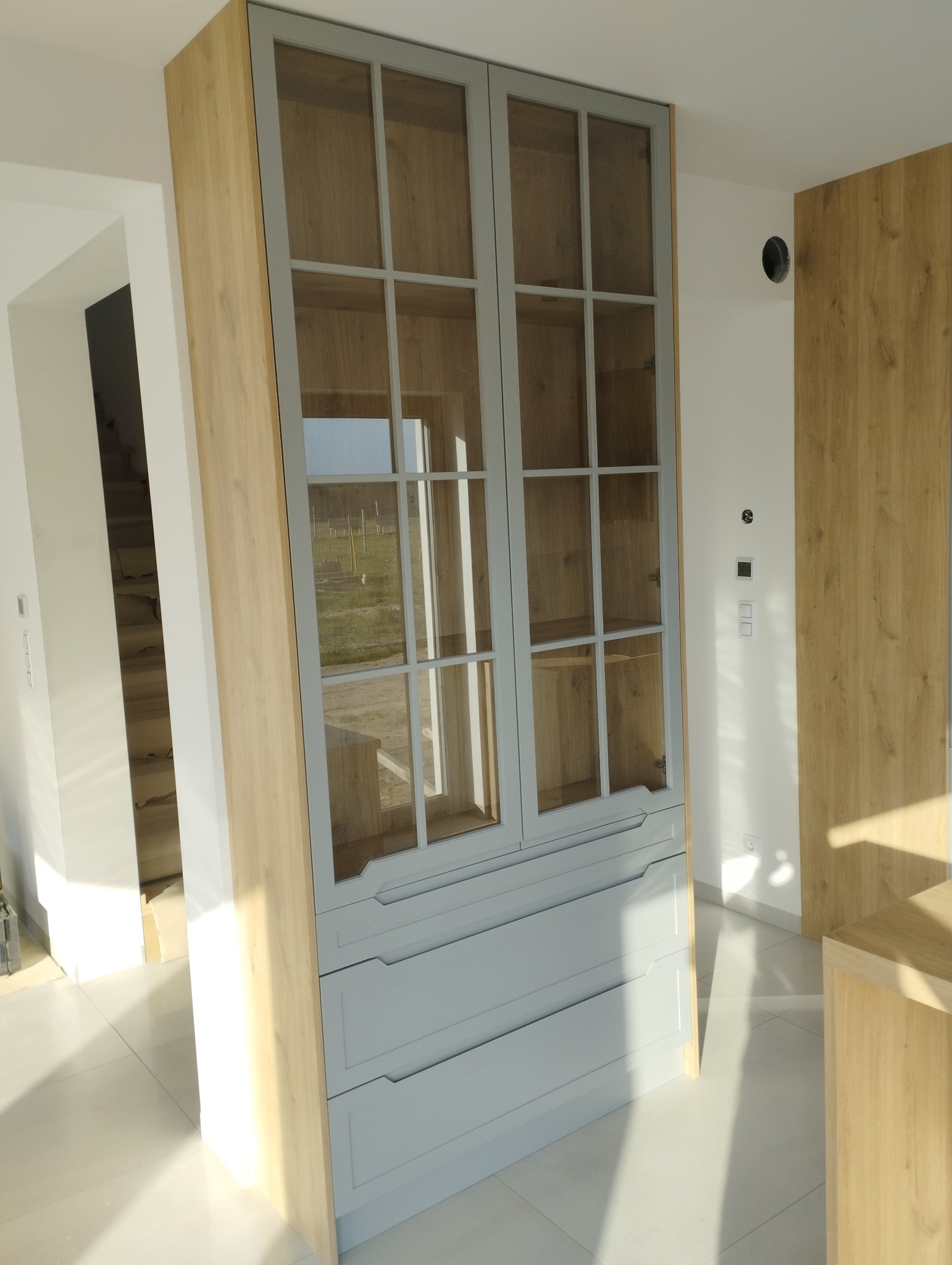 Dwudrzwiowa przeszklona szafa z szufladami w kolorze szarym i drewnianym wykończeniem, widoczna w jasnym wnętrzu z kafelkami na podłodze i schodami w tle.
