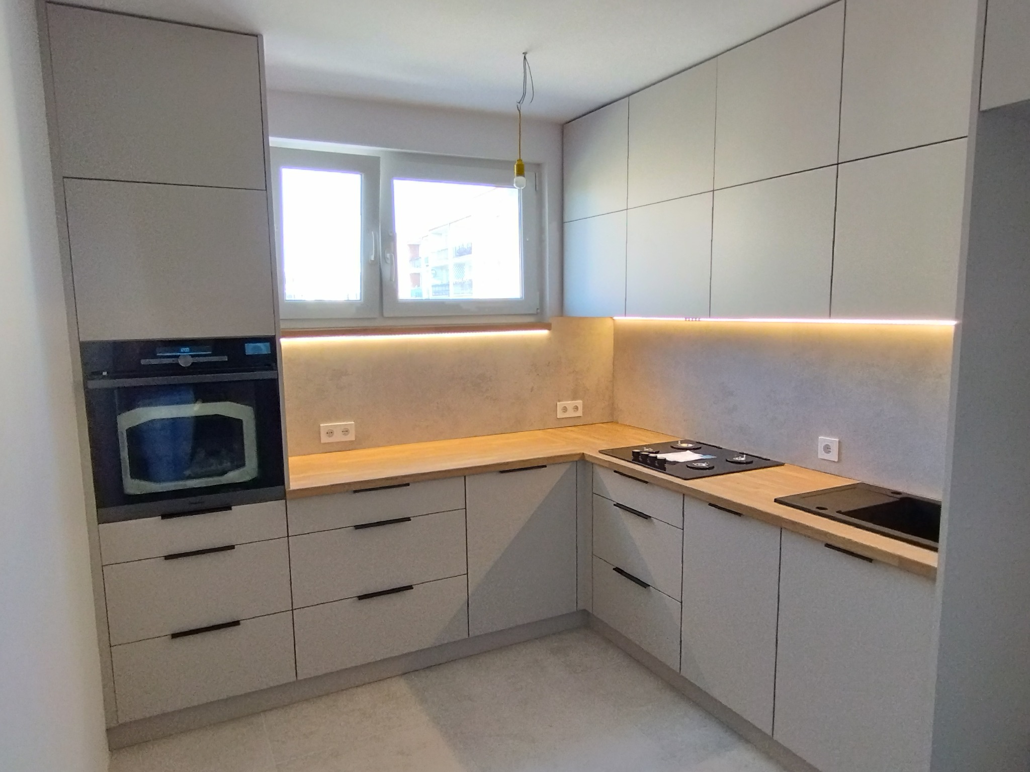 Nowoczesna kuchnia z jasnymi, gładkimi frontami szafek, drewnianym blatem i wbudowanym piekarnikiem, oświetlona listwą LED pod szafkami wiszącymi, z widoczną żarówką zwisającą z sufitu.