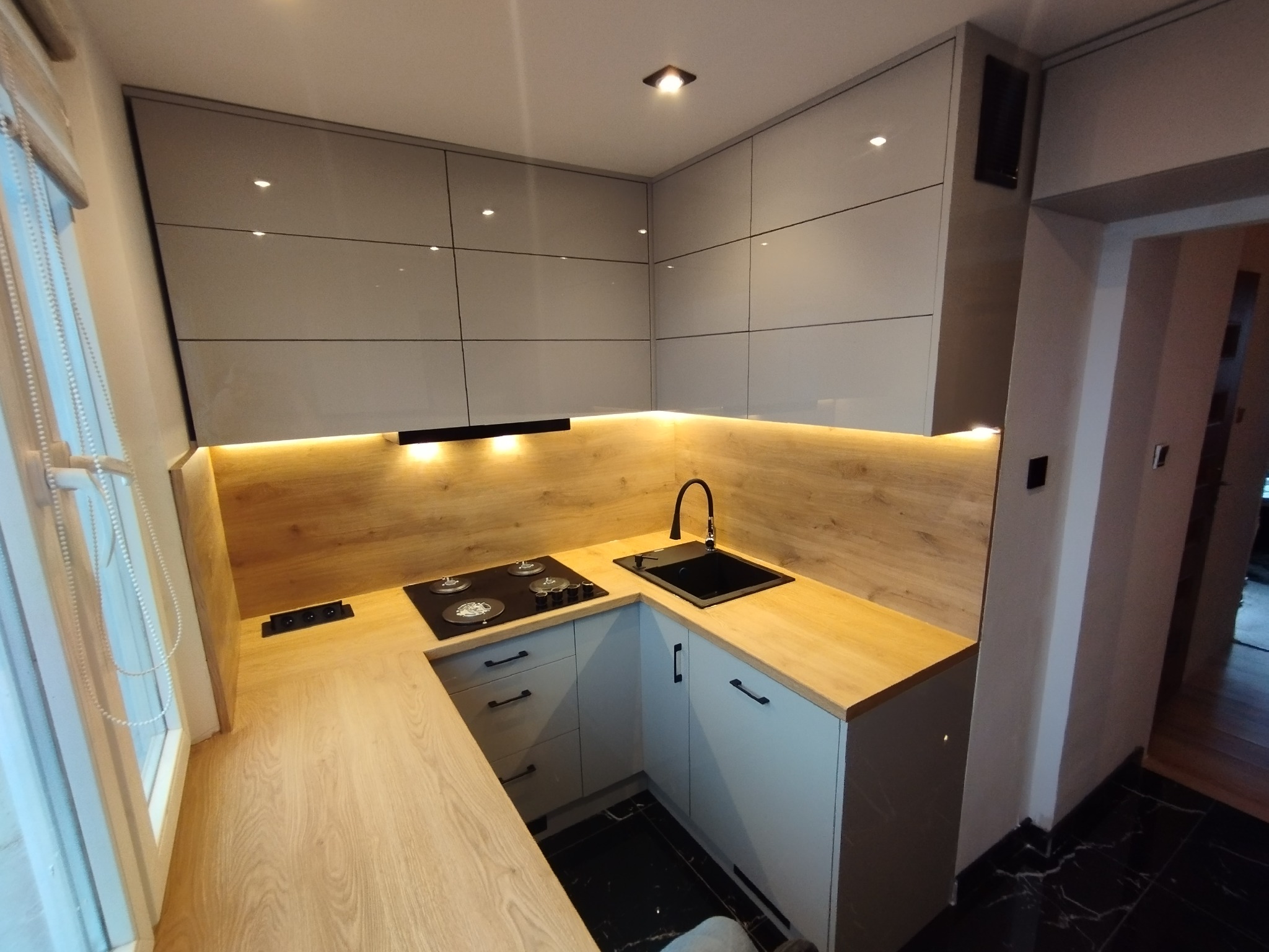 Nowoczesna, jasna kuchnia z drewnianymi blatami i szarymi, błyszczącymi szafkami, oświetleniem LED pod szafkami wiszącymi i czarną armaturą, widoczna płyta grzewcza i gniazdka elektryczne w blacie.