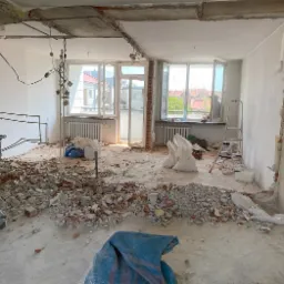 Wnętrze mieszkania w trakcie remontu: usunięte ściany, gruz na podłodze, widoczne instalacje, w tle otwarte okno z widokiem na budynki, w rogu wózek sklepowy.