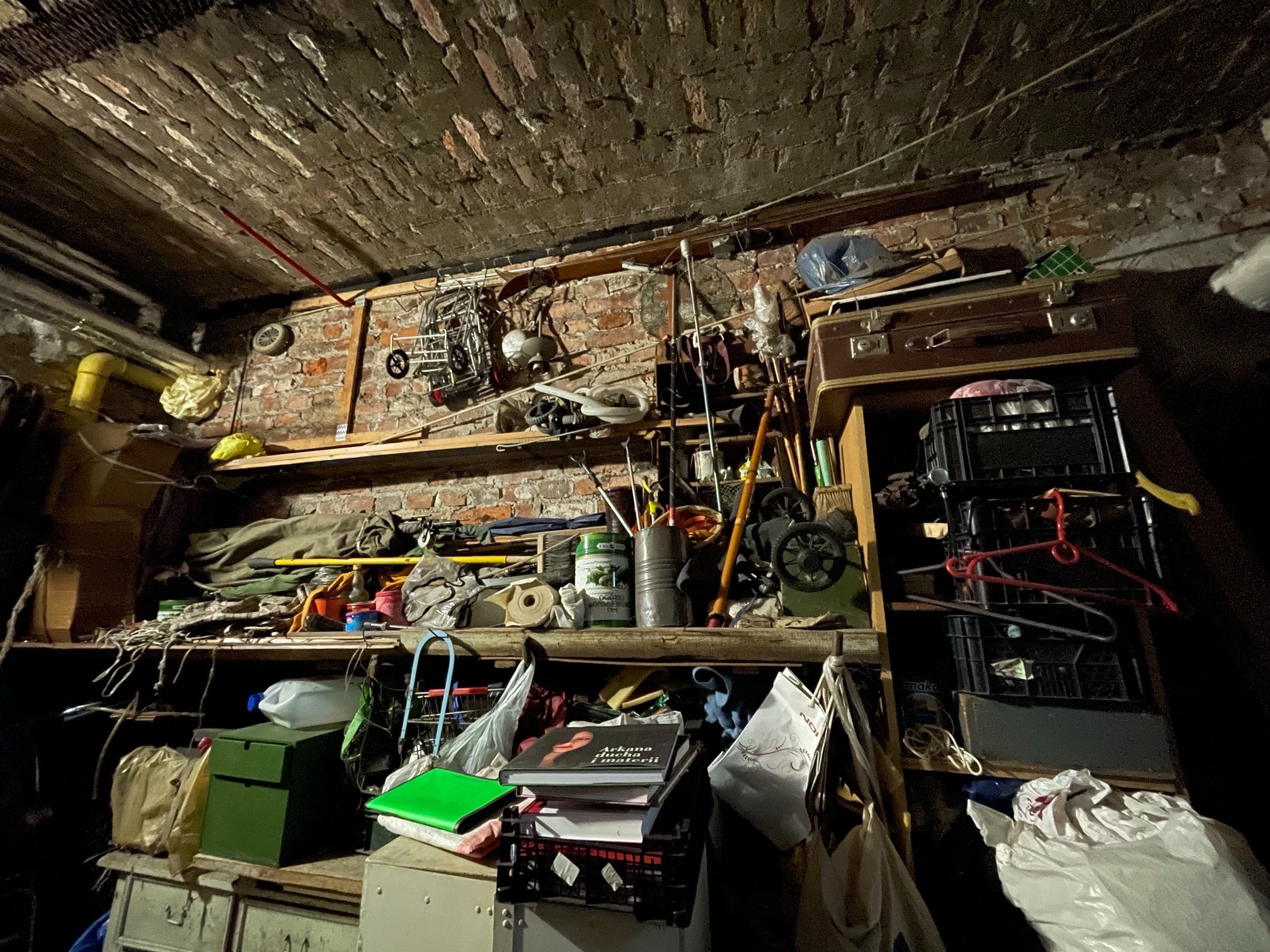 Zastawiony stary strych z ceglanym sklepieniem, wypełniony różnorodnymi przedmiotami na drewnianych półkach, w tym walizki, narzędzia, książki i pojemniki, oświetlony ciepłym światłem.