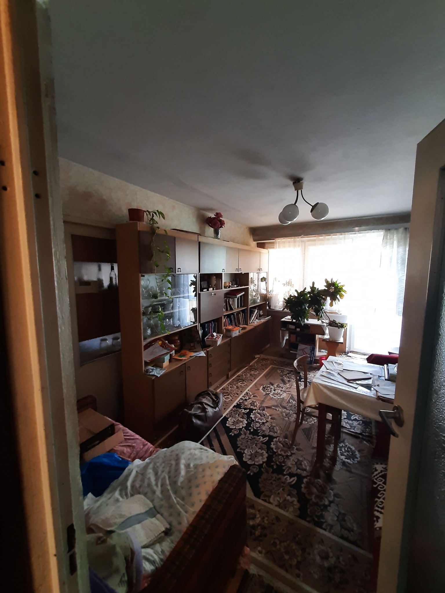 Widok z korytarza na pokój z meblościanką, stołem przy oknie i rozłożonym łóżkiem, w tle jasne okno z firanką.