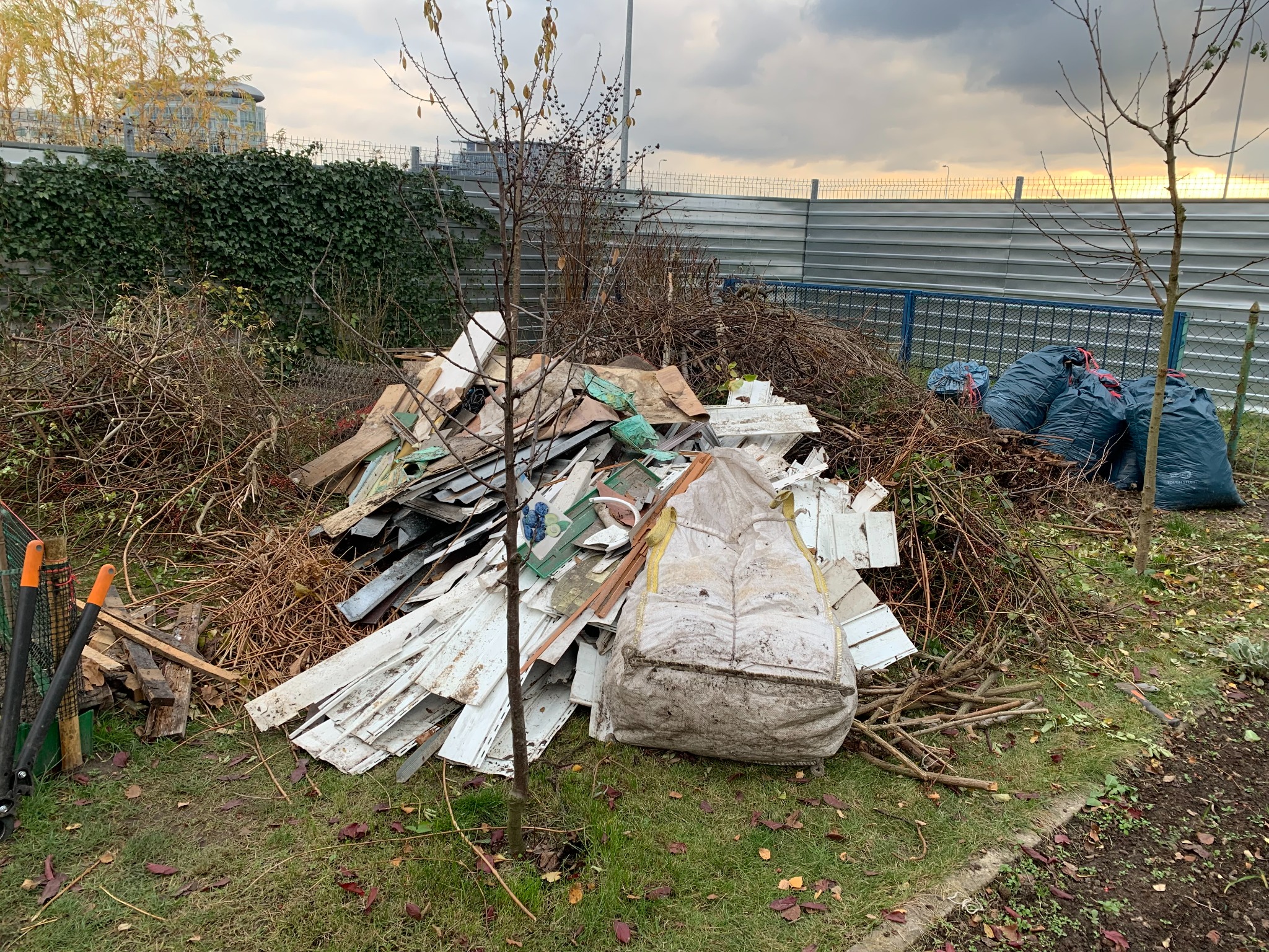 Sterta gruzu, gałęzi i odpadów budowlanych na trawie, obok narzędzi ogrodniczych i worków, z metalowym ogrodzeniem i budynkiem w tle.