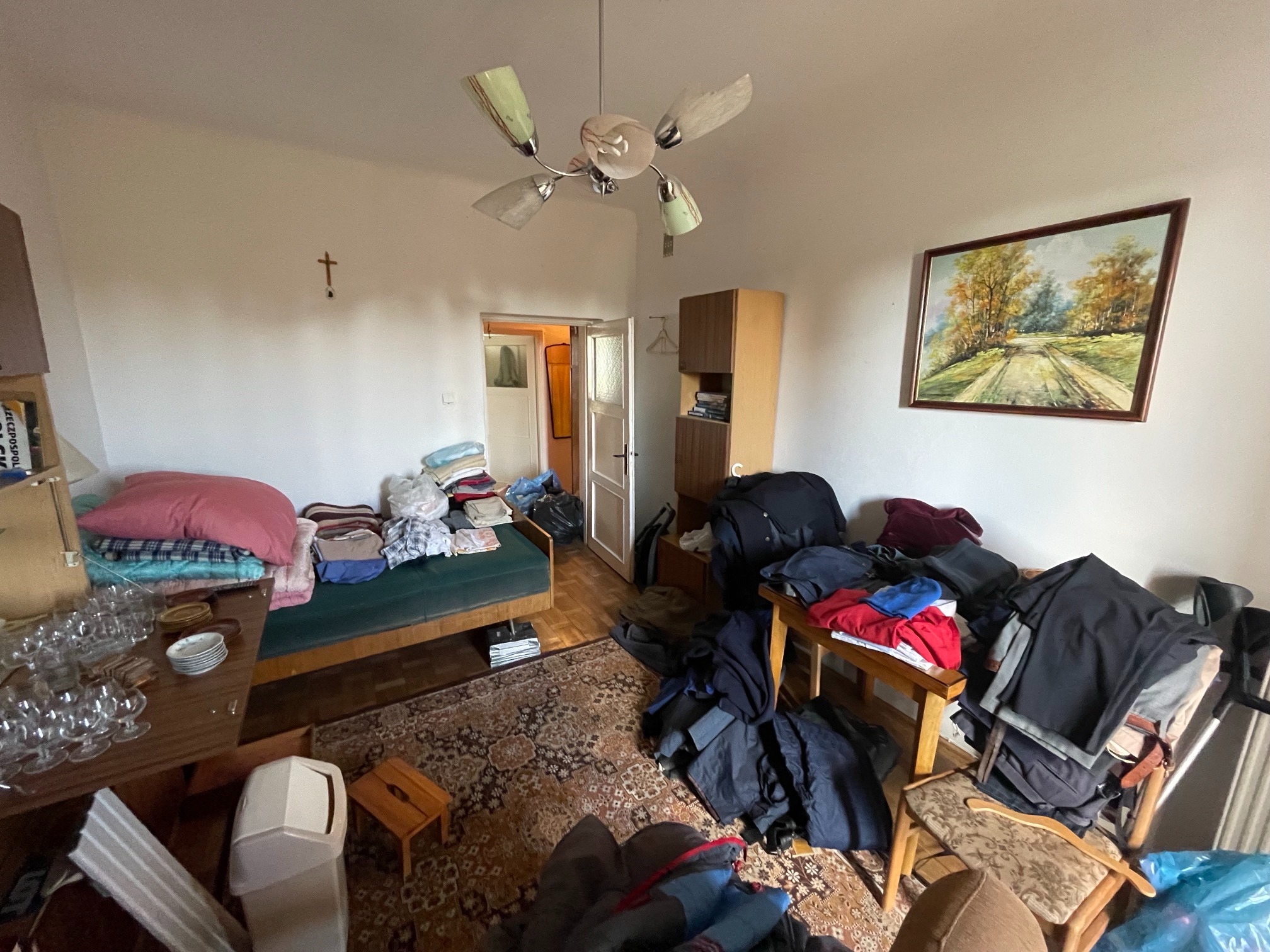 Pokój przed generalnym sprzątaniem: chaotycznie ułożone ubrania na meblach, sterty rzeczy na łóżku i podłodze, naczynia na stole, widoczny dywan z wzorem orientalnym i lampa sufitowa w stylu retro.