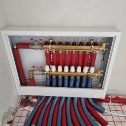 Rozdzielacz ogrzewania podłogowego w białej skrzynce, widoczne czerwone i niebieskie rury wychodzące na folię izolacyjną z siatką.