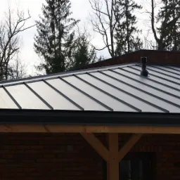 Dach pokryty panelami na rąbek stojący w kolorze grafitowym, widoczny kominek wentylacyjny i fragment drewnianej konstrukcji budynku z ceglaną ścianą w tle.