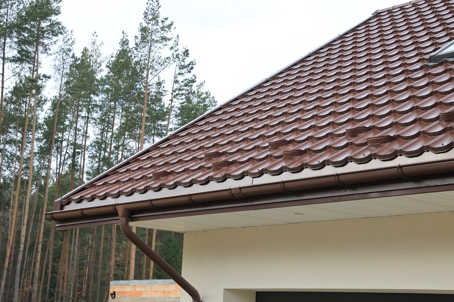 Brązowa blachodachówka Armano na dachu domu jednorodzinnego z zamontowanymi rynnami w kolorze dachu, na tle lasu iglastego.