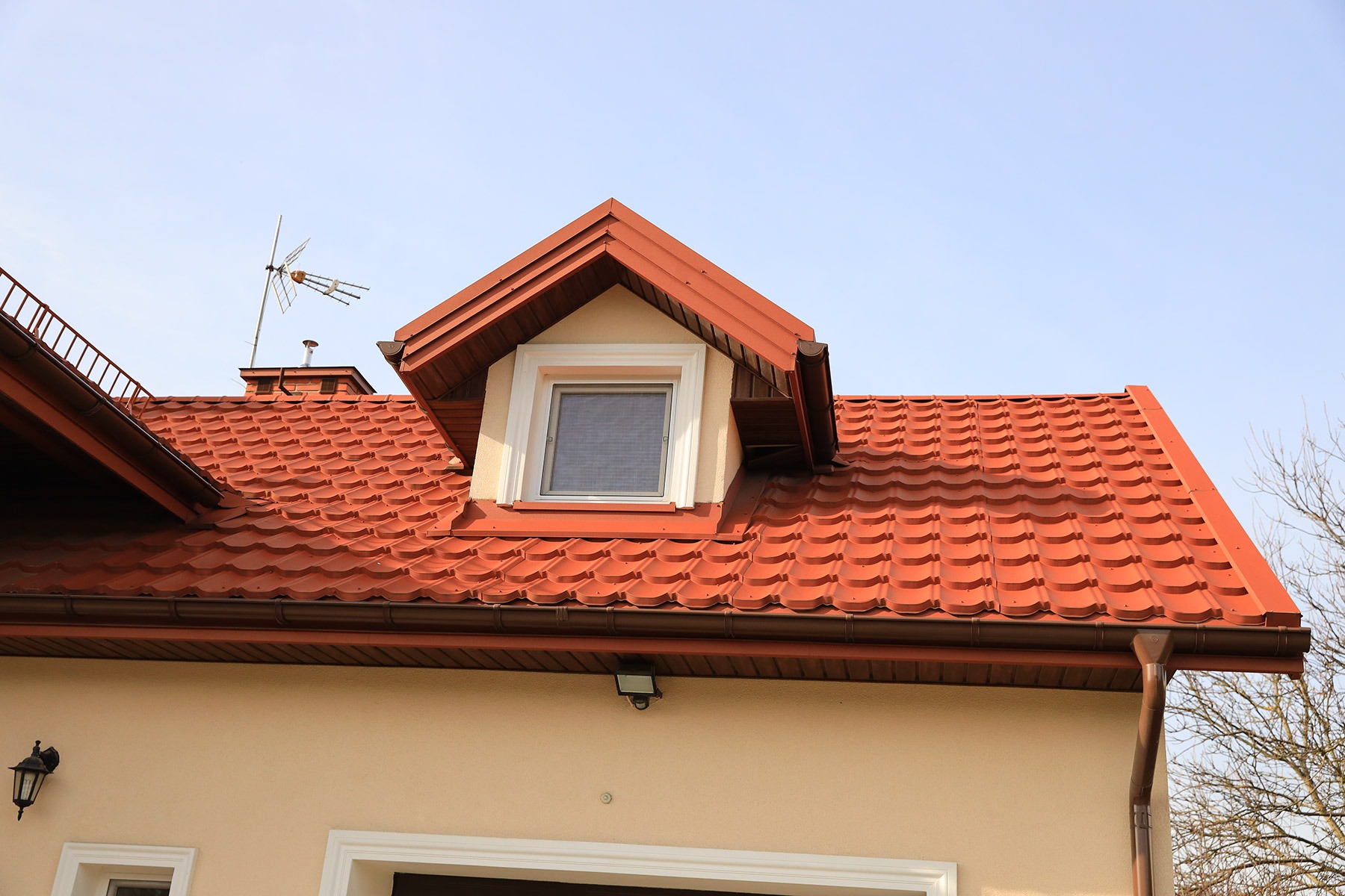 Czerwona blachodachówka Armano na dachu domu z oknem mansardowym i brązowymi rynnami, widoczna antena na kominie.