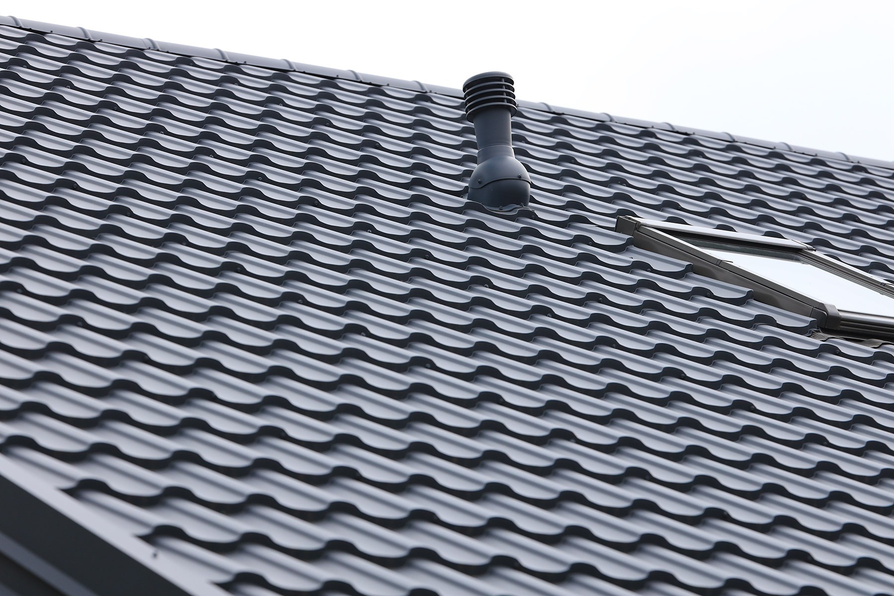 Szary dach pokryty blachodachówką Barcelona z kominkiem wentylacyjnym i oknem dachowym, widoczne regularne fale blachy.