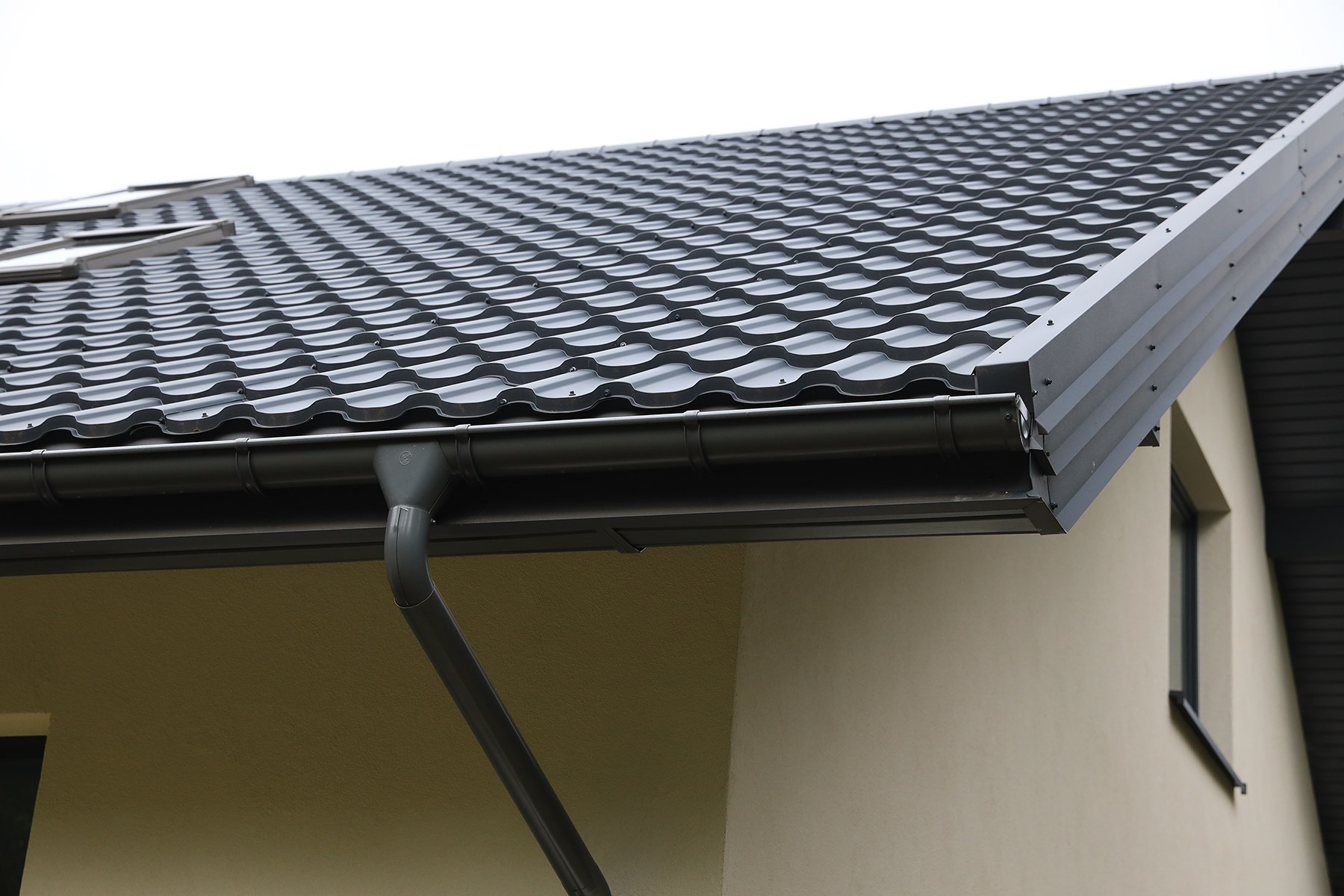 Ciemnoszara blachodachówka Barcelona na dachu domu z zamontowanymi rynnami w tym samym kolorze, widok na fragment dachu z oknami dachowymi i narożnik budynku.