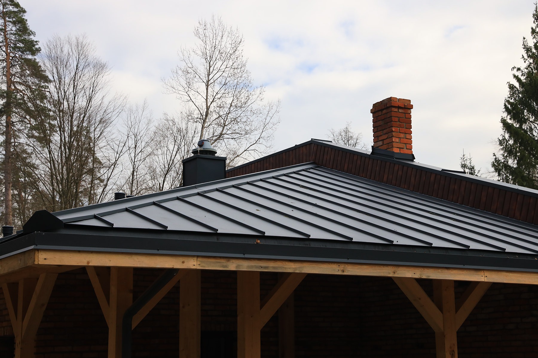 Czarny panel na rąbek stojący na dachu z kominem z cegły, widok z poziomu drewnianej konstrukcji z ceglanym wypełnieniem.