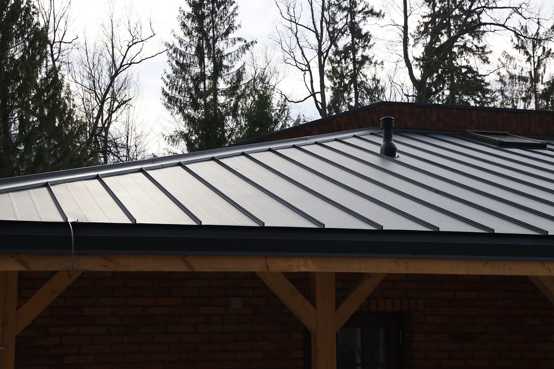 Dach pokryty panelami na rąbek stojący w kolorze grafitowym, widoczny kominek wentylacyjny i fragment drewnianej konstrukcji budynku z ceglaną ścianą w tle.
