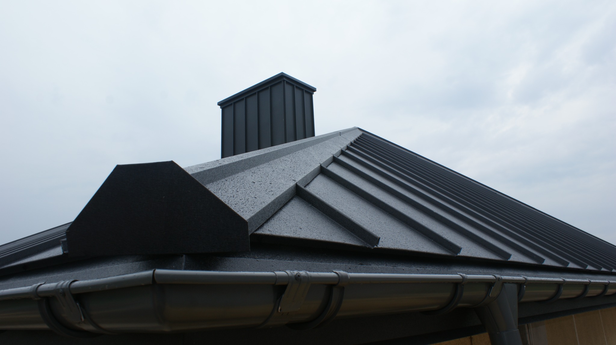Nowoczesny dach z paneli na rąbek stojący, widoczne orynnowanie w kolorze grafitowym i komin wentylacyjny, całość w pochmurny dzień.