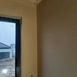 Świeżo nałożone tynki na ścianach wewnętrznych pomieszczenia, widoczny fragment okna z widokiem na dom i błotniste podwórko, w rogu widoczny fragment drabiny malarskiej.