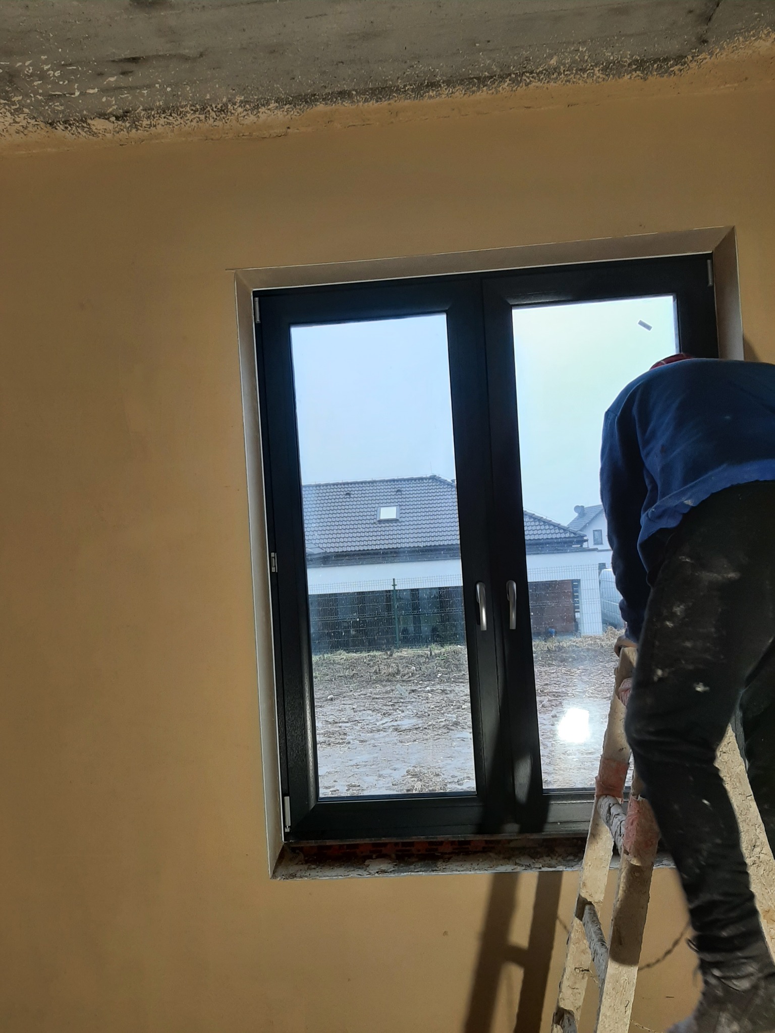 Pracownik na drabinie, w trakcie tynkowania ściany wokół okna w surowym stanie; widoczny fragment sufitu i okno z widokiem na dom.