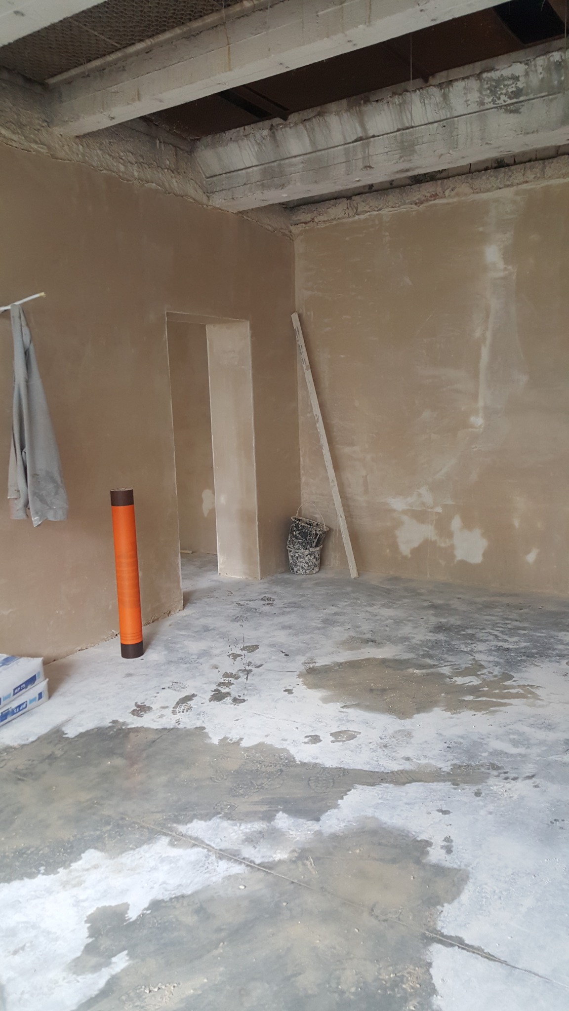 Pokój w trakcie tynkowania maszynowego: świeżo nałożony tynk na ścianach, mokra posadzka, widoczne worki z zaprawą, pomarańczowa tuba i wiadro.