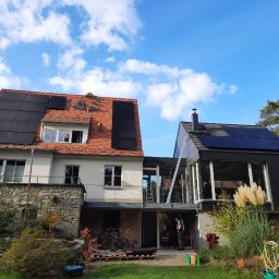 ProMat - Dwa budynki z panelami słonecznymi na dachach, jeden z czerwoną dachówką, drugi nowoczesny z czarną elewacją, drabina oparta o budynek, widok z ogrodu.