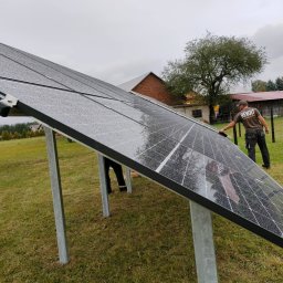 ProMat - Instalacja paneli fotowoltaicznych na metalowych podporach na trawiastym terenie, pracownik w koszulce z logo firmy, widoczne krople deszczu na panelach.