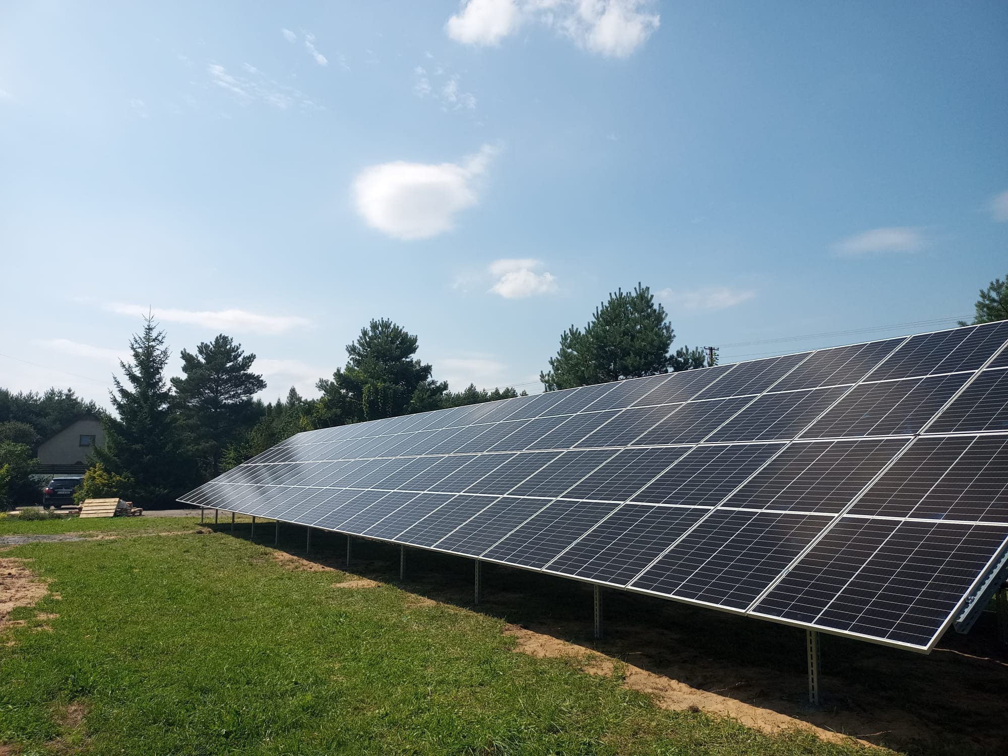 Rząd paneli słonecznych na metalowych wspornikach na trawiastym terenie, drzewa w tle, błękitne niebo z pojedynczymi chmurami.