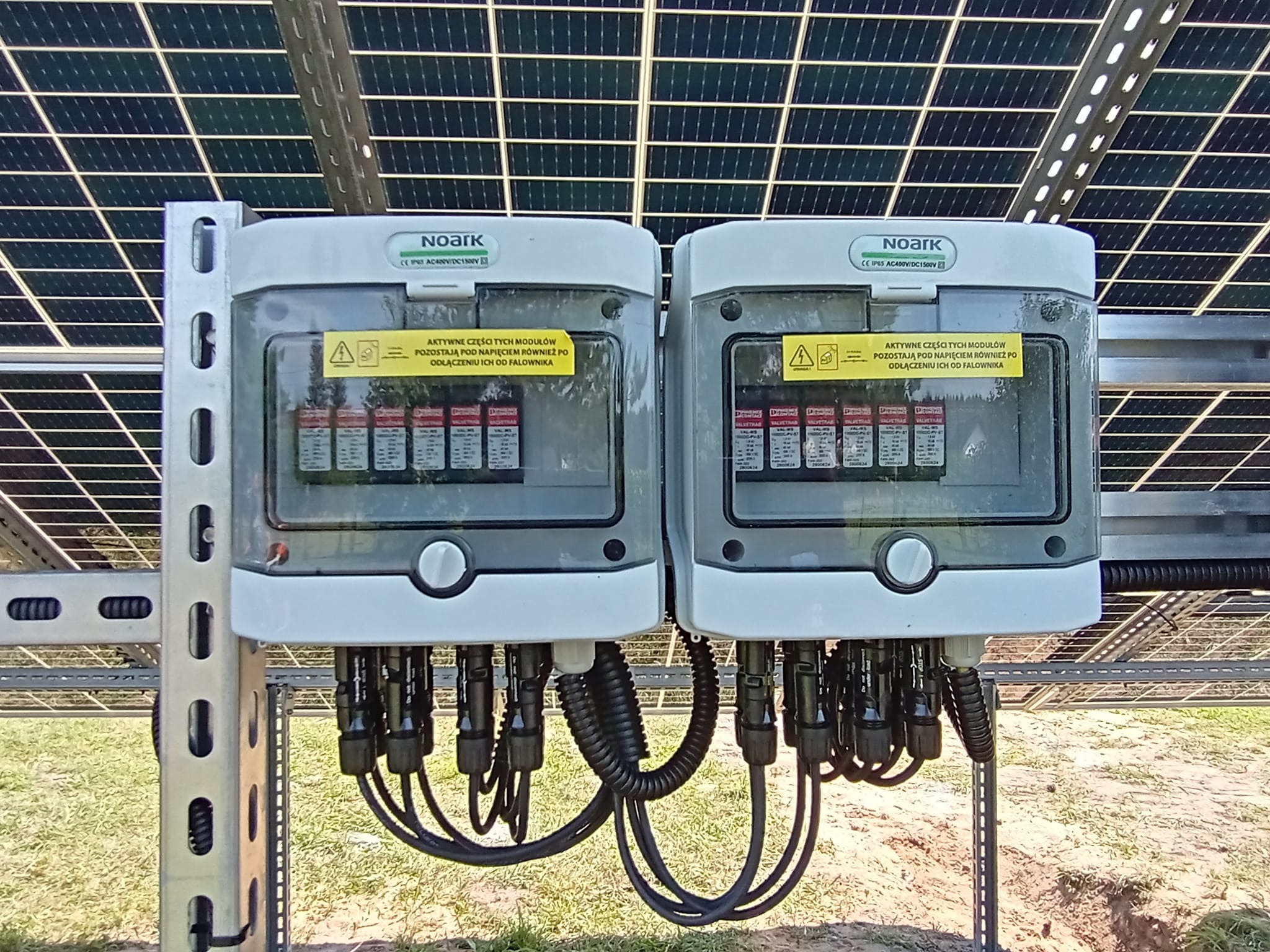 Dwa połączone białe moduły przyłączeniowe firmy Noark, z widocznymi kablami i ostrzeżeniami o napięciu, zamontowane na metalowej konstrukcji pod panelami słonecznymi.