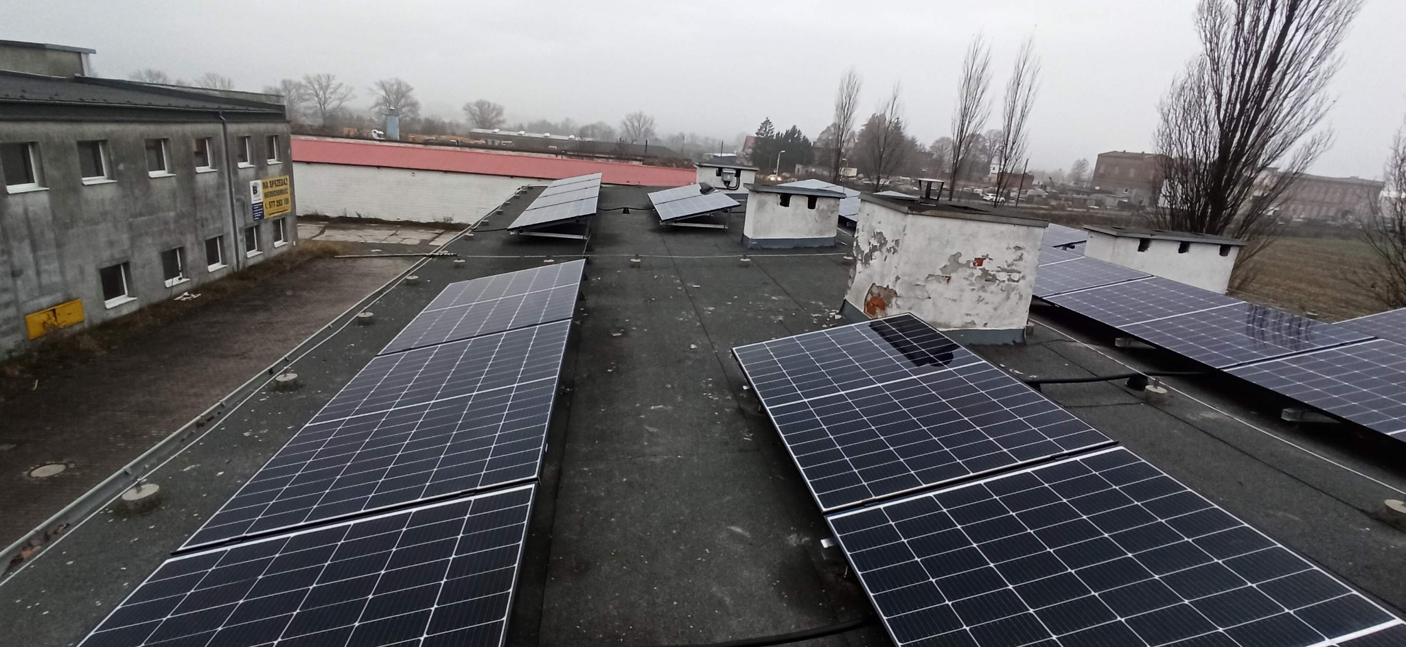 Instalacja paneli słonecznych na płaskim dachu budynku przemysłowego w pochmurny dzień, widoczne kominy wentylacyjne i budynki w tle.