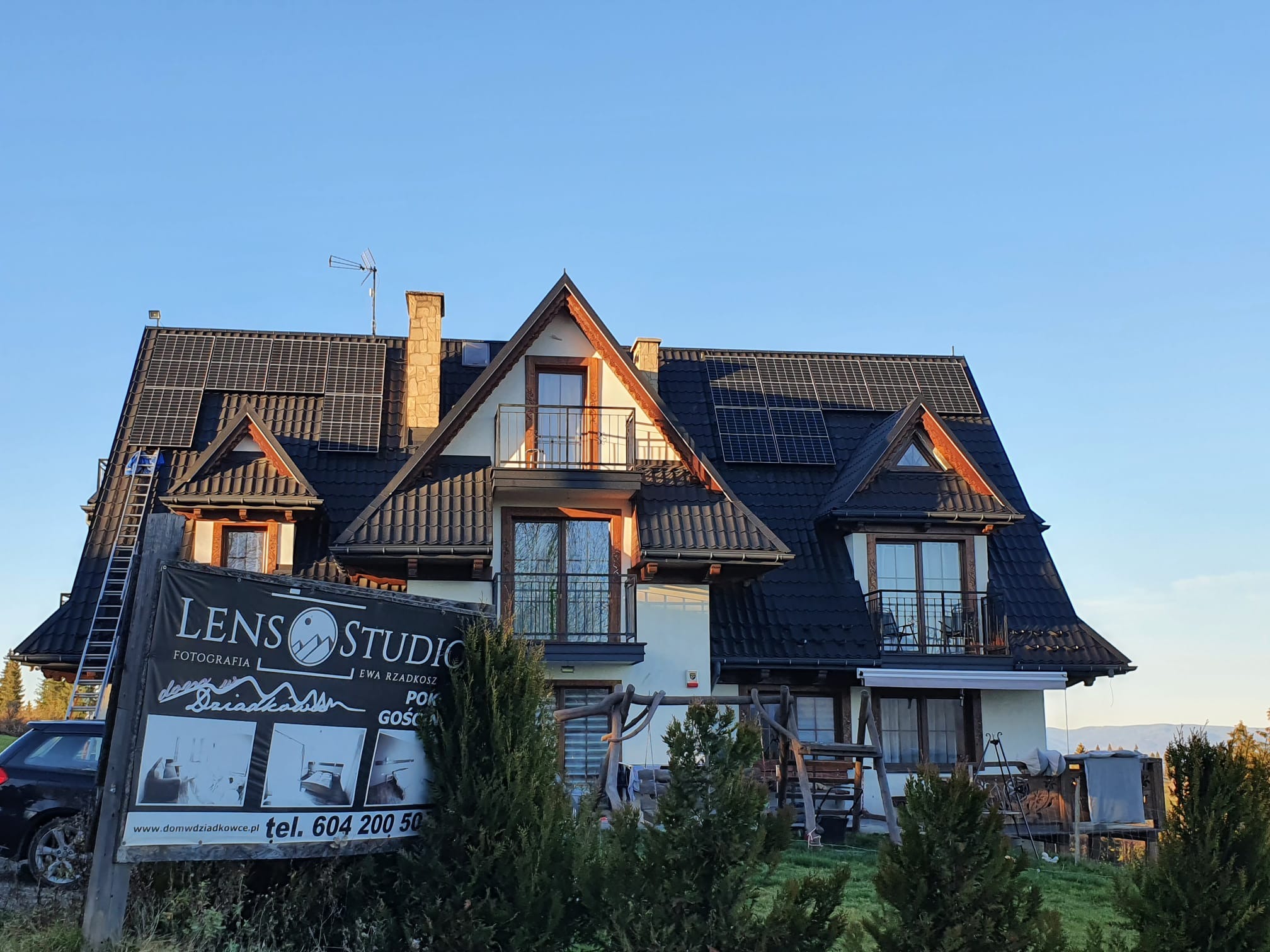 Dom z ciemnym dachem, na którym zamontowane są panele słoneczne, widoczna drabina oparta o dach i baner reklamowy Lens Studio z przodu.