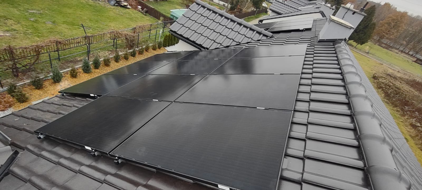 Czarny panel słoneczny na ciemnym dachu z dachówki ceramicznej, widok z góry na dom z ogrodem w tle.