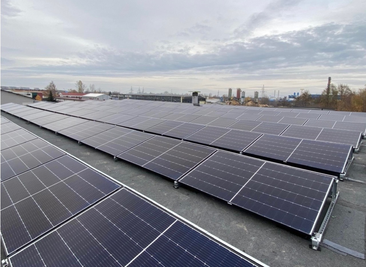 Rząd paneli słonecznych na płaskim dachu budynku przemysłowego, widok z góry przy pochmurnym niebie.