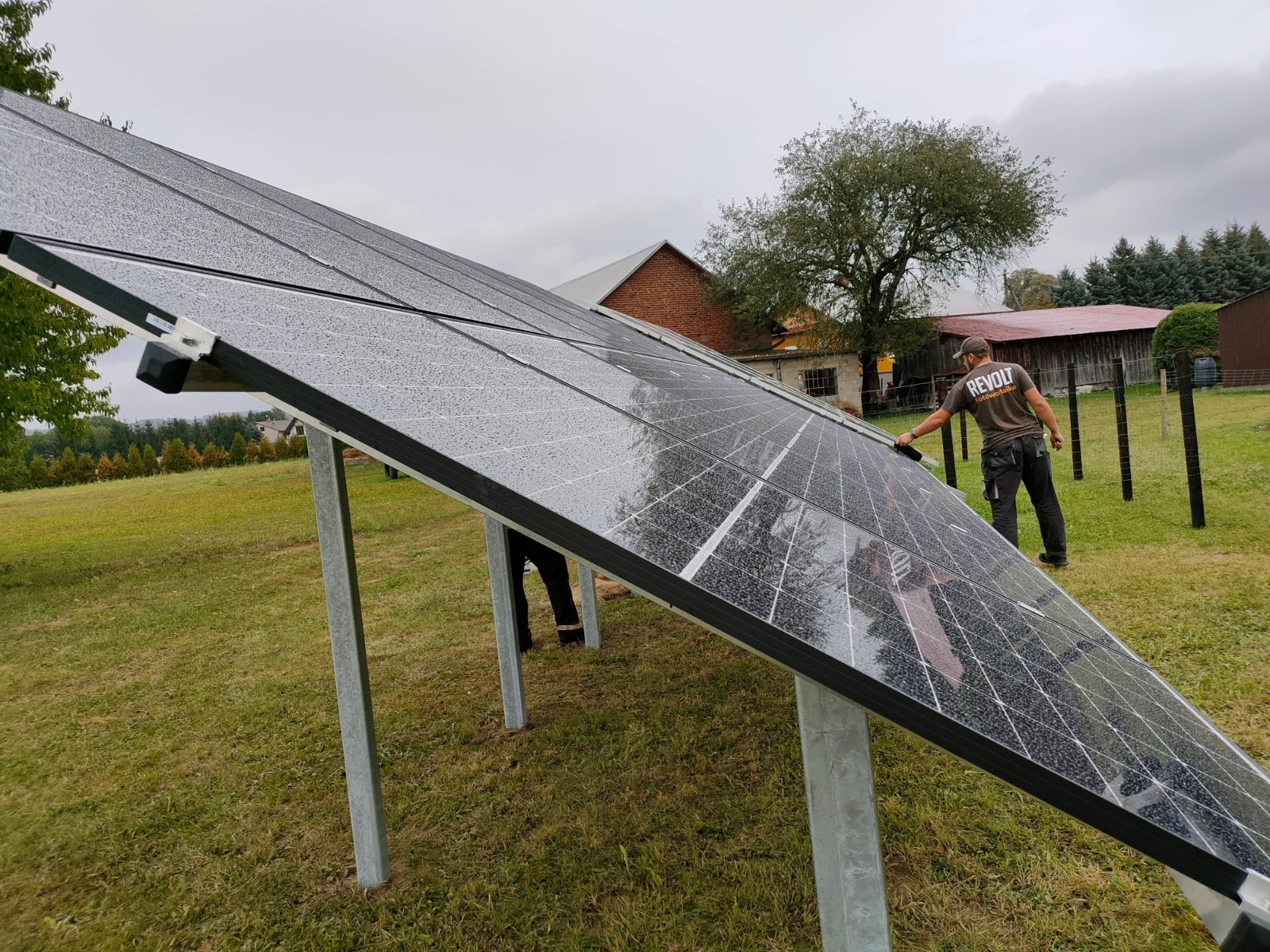 Instalacja paneli fotowoltaicznych na metalowych podporach na trawiastym terenie, pracownik w koszulce z logo firmy, widoczne krople deszczu na panelach.